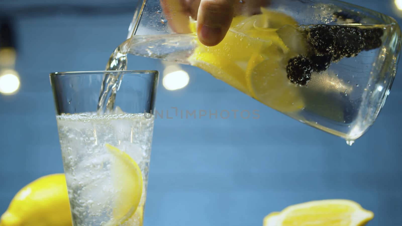 Extreme close up pouring lemonade with dewberry and lemon into a glass. Blurry lamps on blue background