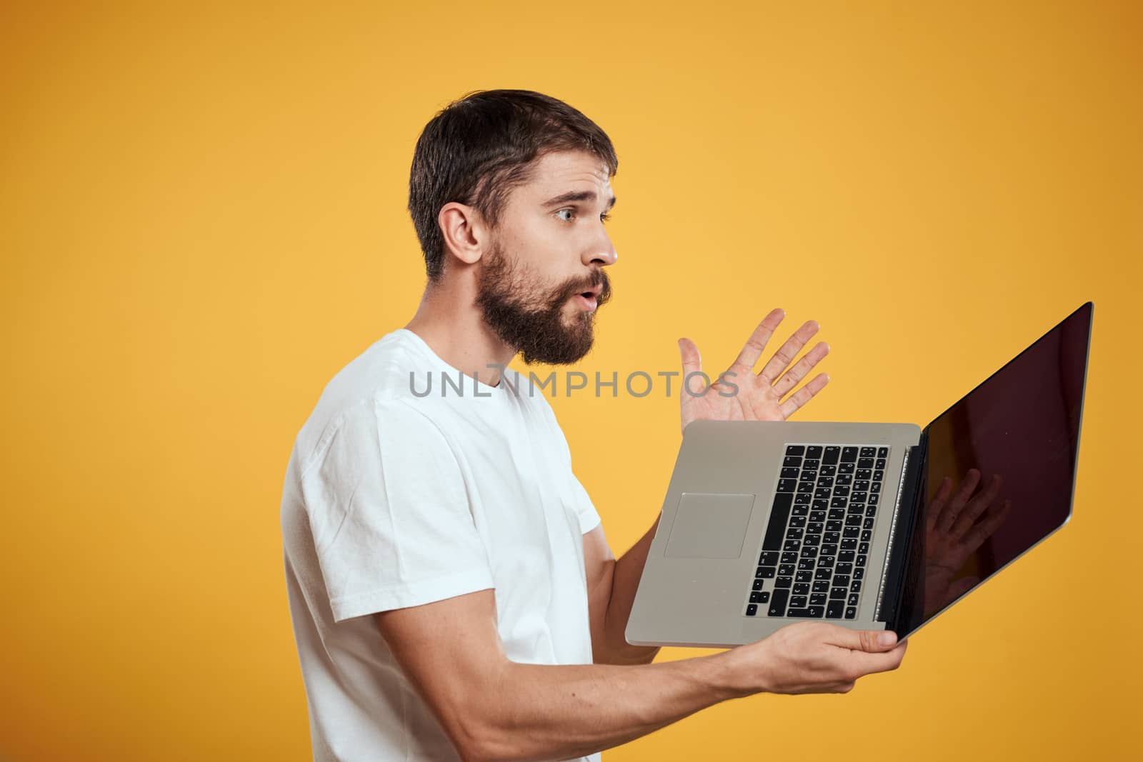 A man with a beard holds a laptop in his hand on a yellow background keyboard monitor new technologies side view by SHOTPRIME