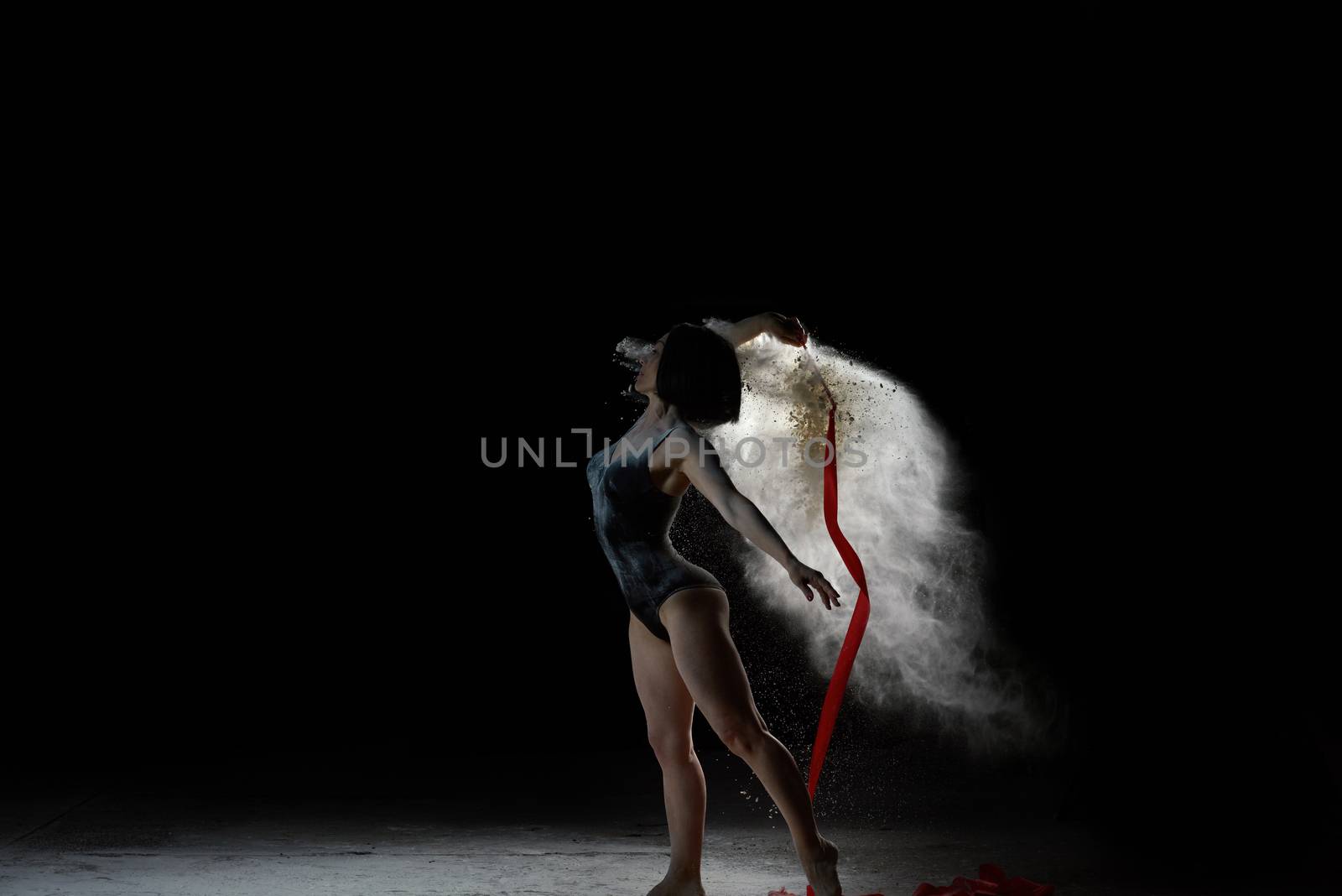 beautiful caucasian woman in a black bodysuit with a sports figure dancing with red ribbon on a black background, particles of flour fly in different directions