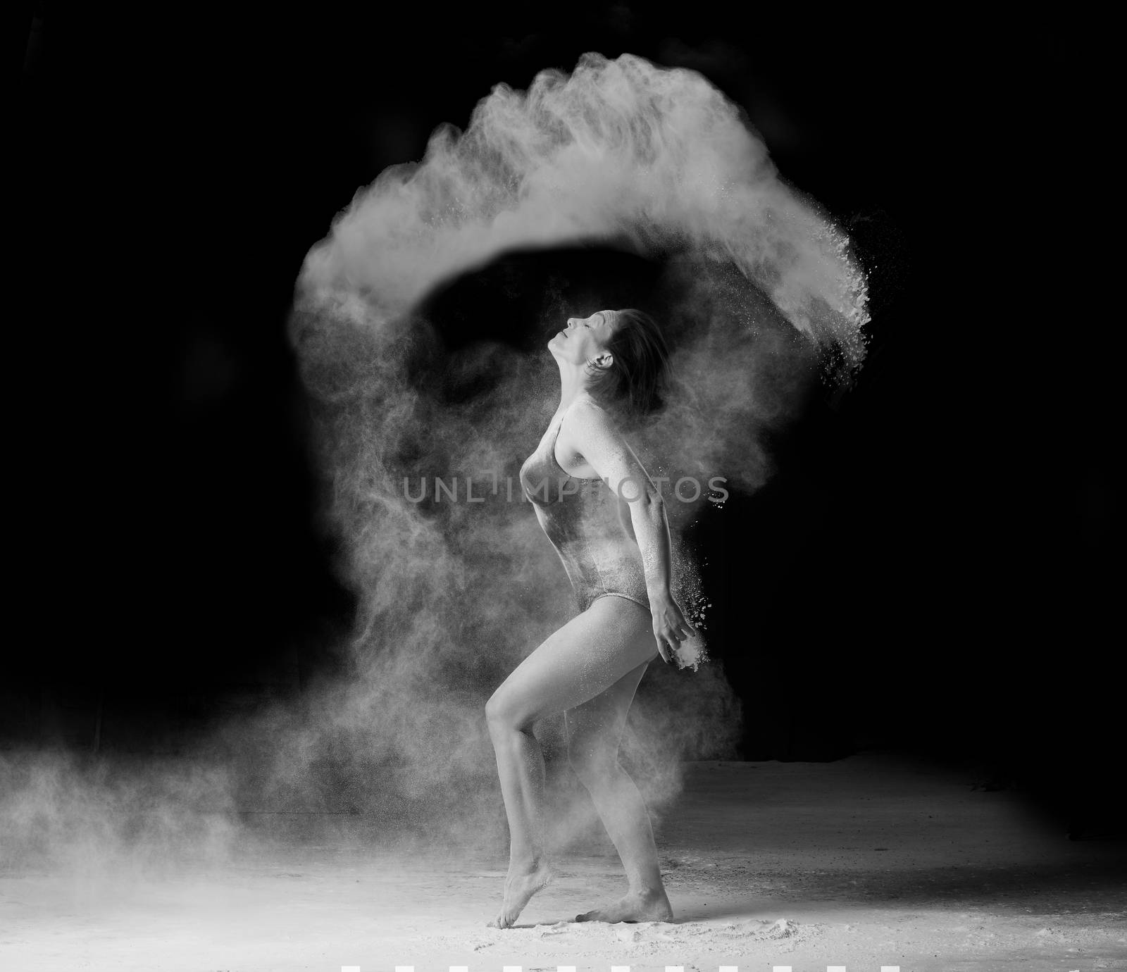 beautiful caucasian woman in a black bodysuit with a sports figure is dancing in a white cloud of flour on a black background, black and white toning