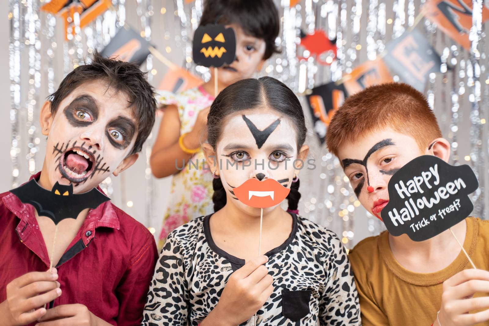 Group of kids in Halloween costumes making gesticulating scary or spooky faces by holding booth sprops on decorated background by looking into camera