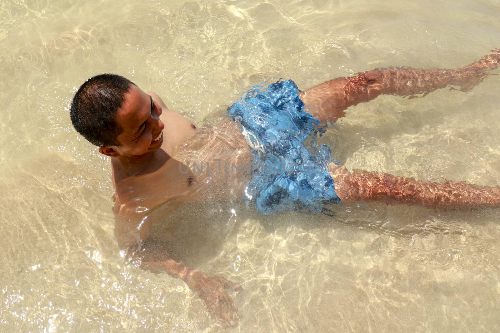 Smiling asian man lying in shallow Water. Sunny Summer Day. Laughing teenager sitting by the sea. Handsome man lying down in the shallow sea.