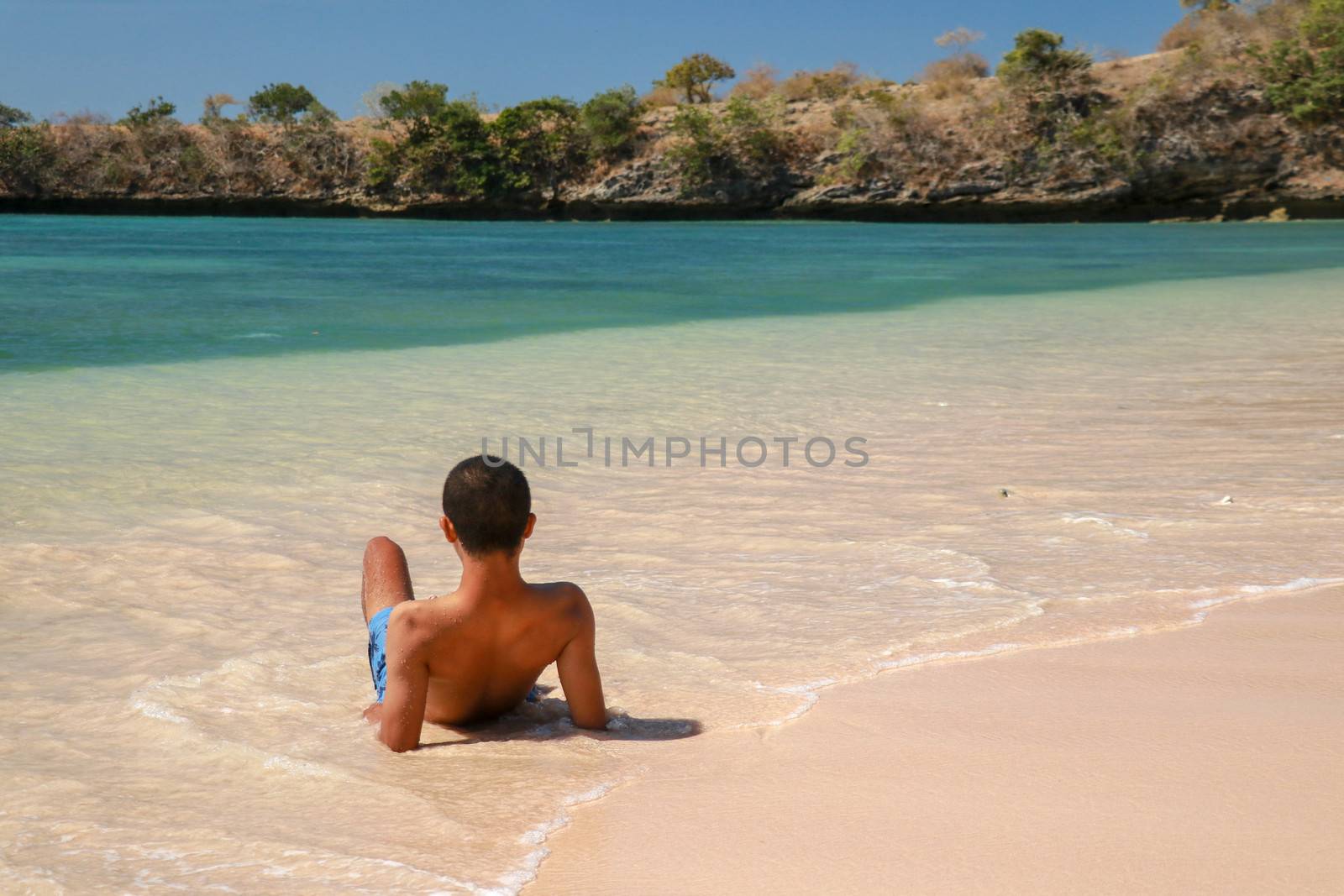 Asian teenager in swimming shorts lying down in calm sea. Man relaxing on a tropical beach. Young tanned man taking sunbath on sand beach. Back view of handsome man in the shallow water.
