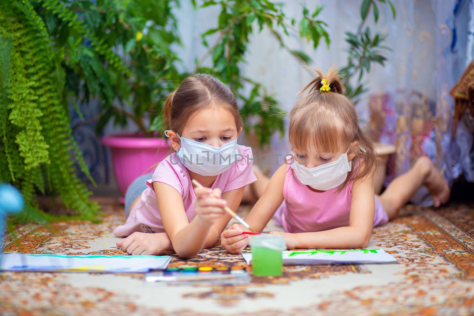 Two girls in protective medical masks paint with paints lying on the floor at home or in kindergarten. Children in self-isolation or quarantine