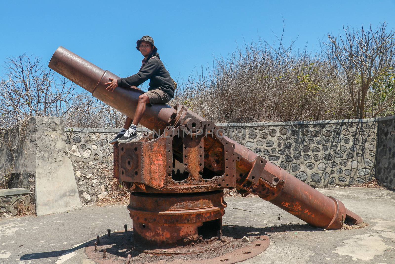 An Asian teenager sits on a historic World War II cannon. Military stone fortifications from World War II. A young guy sitting on an ancient cannon. Lombok, Indonesia.