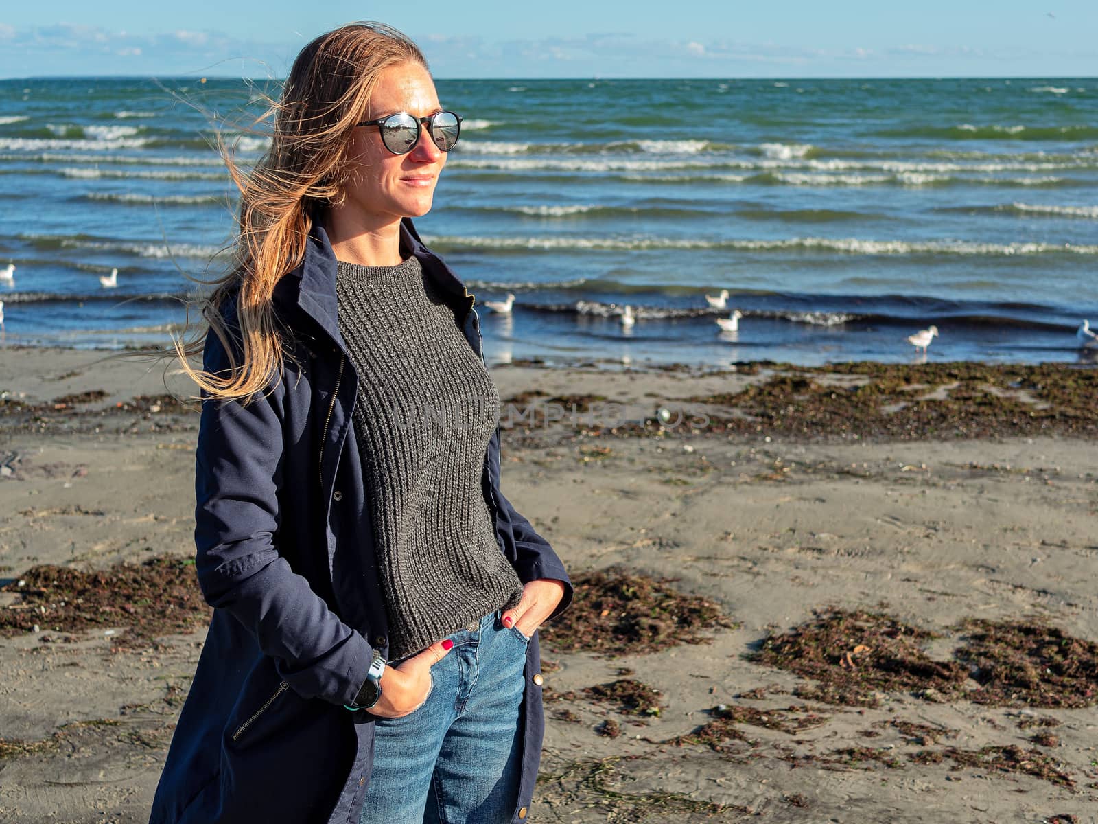 Pretty young girl ashore the Baltic sea. Young woman with long hair and dark coat in nature. autumn beach of the Baltic Sea. Blue sea on the backgroun. Copy space