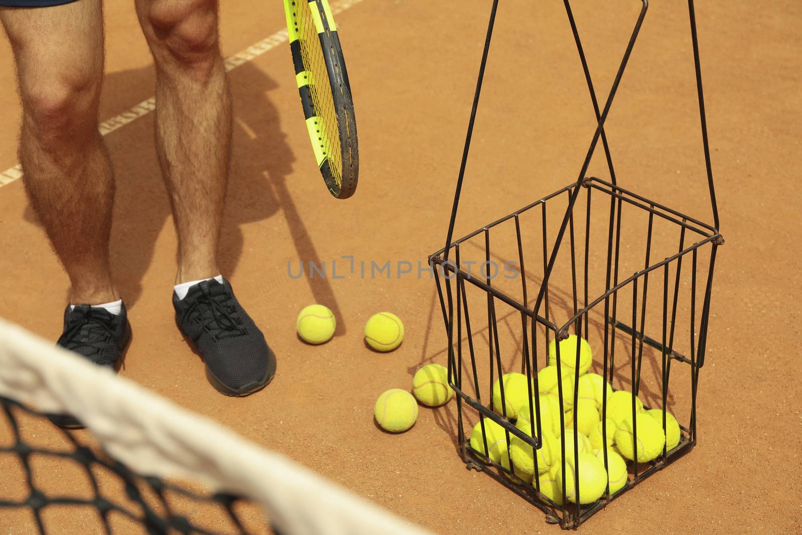 Man hold racket on clay court with basket of tennis balls