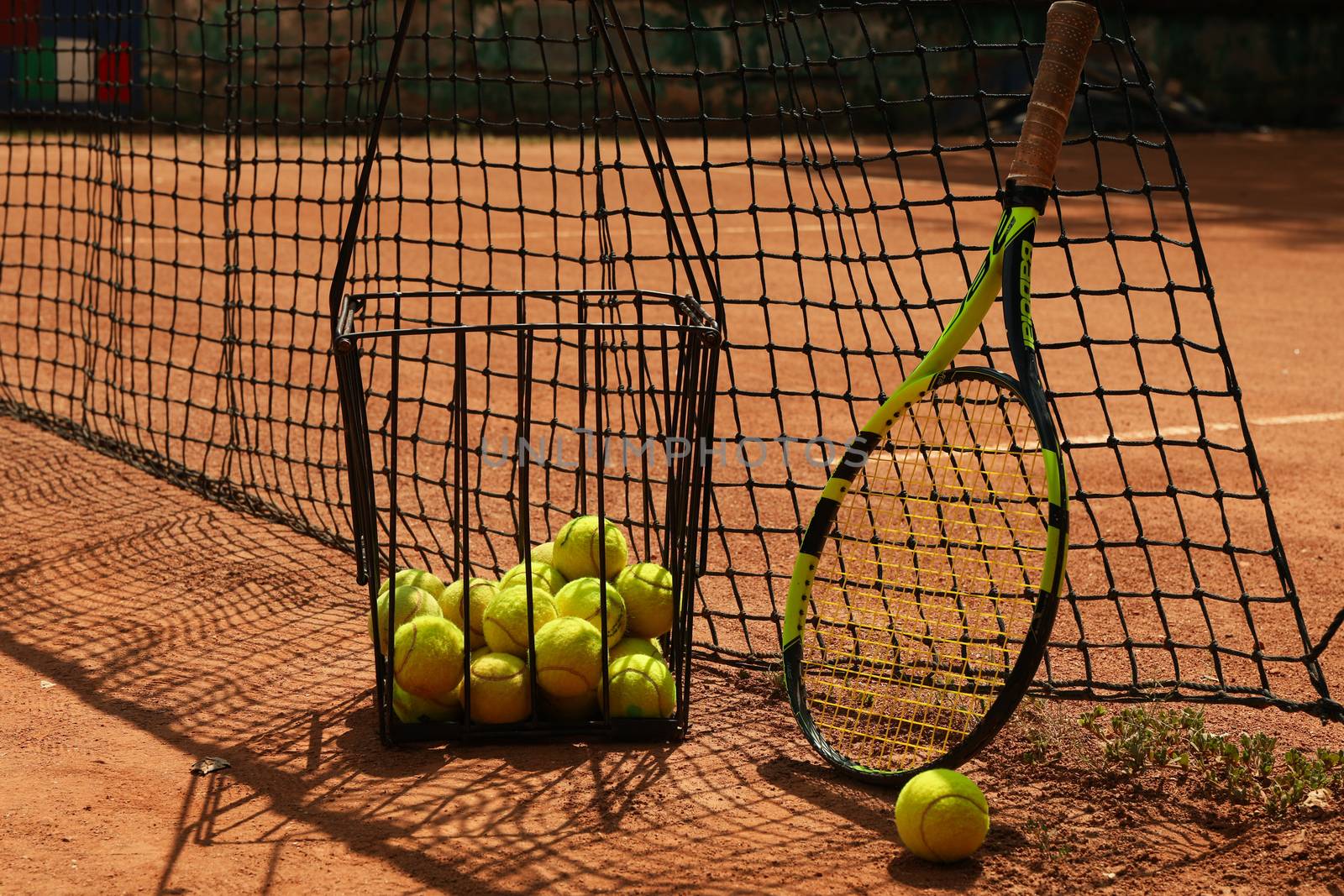 Basket with tennis balls and racket against net on clay court