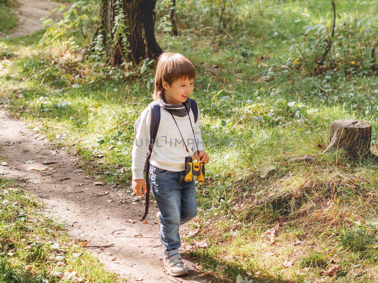 Curious boy is hiking in forest lit by sunlight. Outdoor leisure activity for kids. Child with binoculars and backpack.