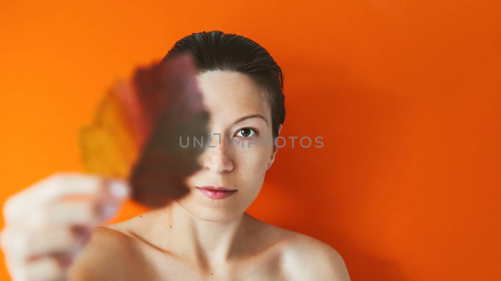 Portrait of Asian woman closing her eye with autumn leaf. Model on orange monochrome background. Natural beauty on bright and colorful backdrop. Banner with copy space.