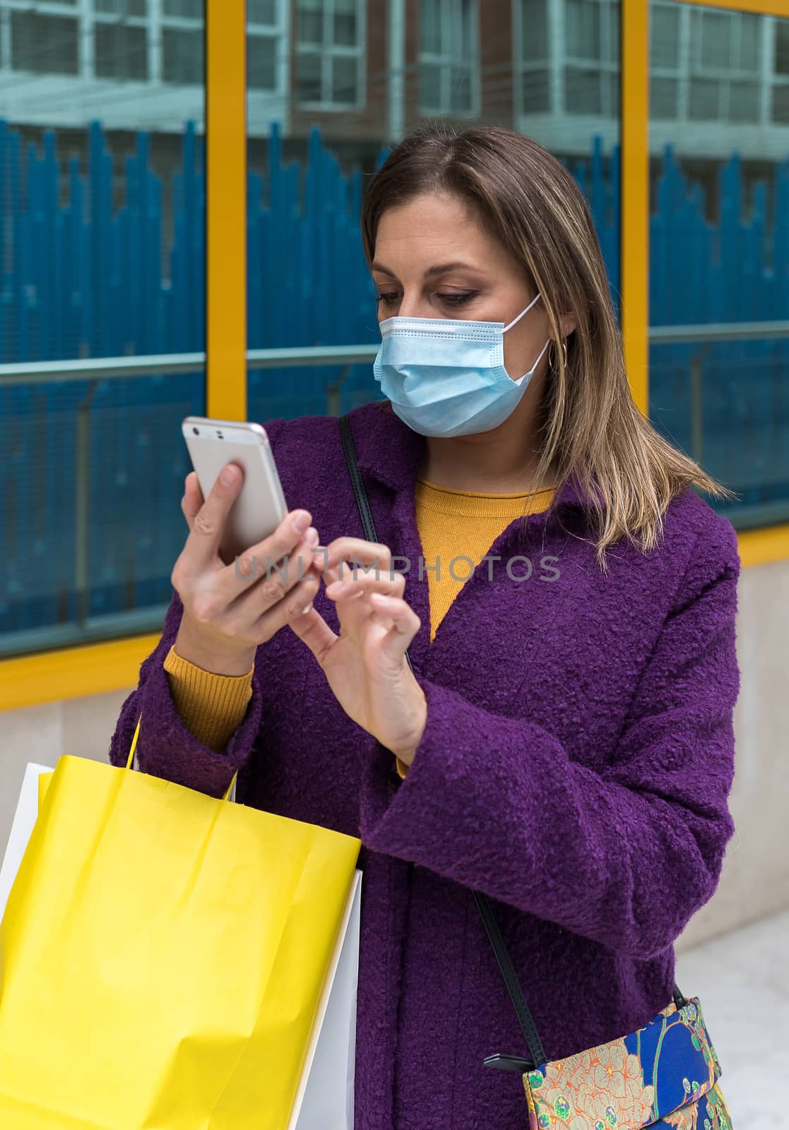 Middle-aged blonde woman with face mask looking at mobile phone while holding shopping bags on her arm