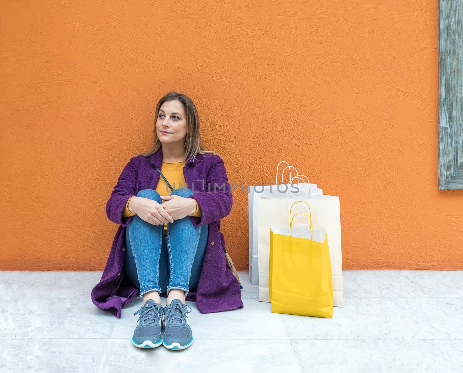 Smiling middle-aged blonde woman sitting on the floor with shopping bags on a orange background