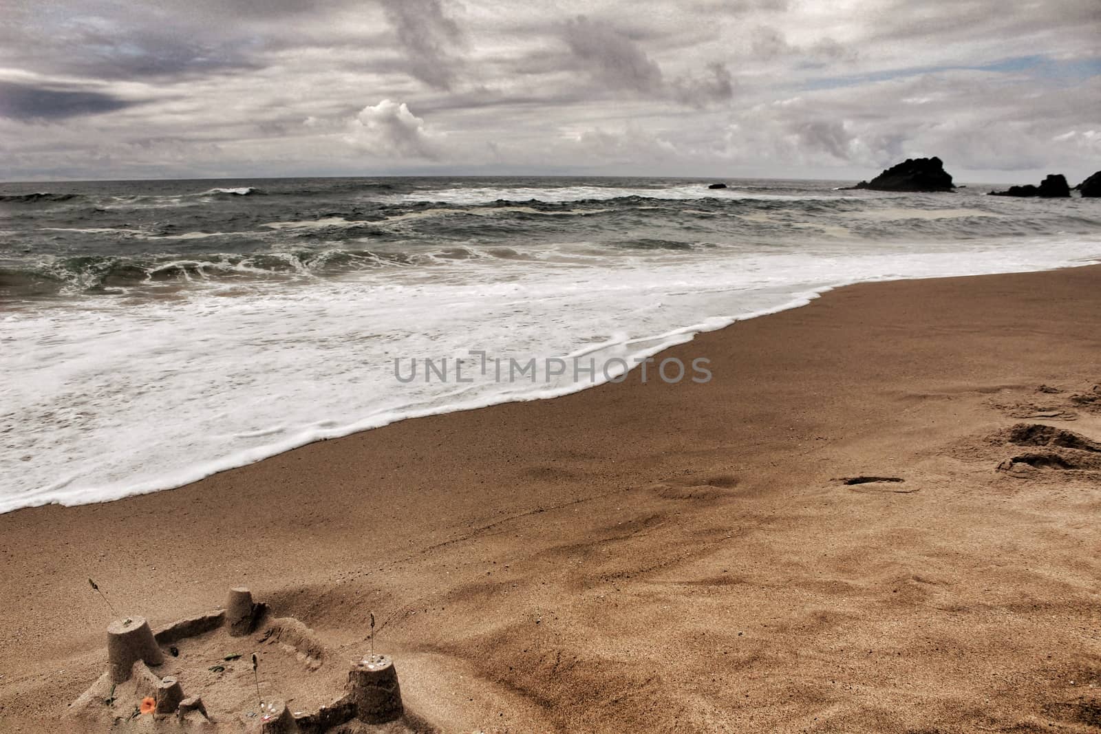 Brave ocean, rock formations and cloudy drama sky on the beach of Adraga in Sintra, Lisbon, Portugal. Sandcastle in the foreground.