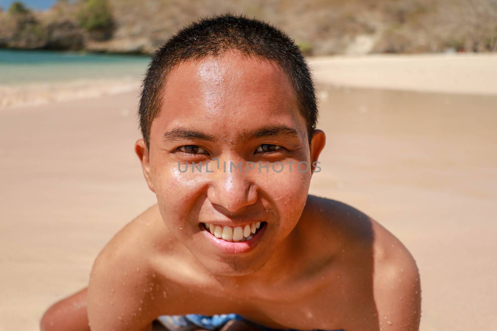 portrait of a model guy lying on his stomach on a blue beach tanned and looking sexually at the camera. Smiling Asian teenager lying on the sandy beach.