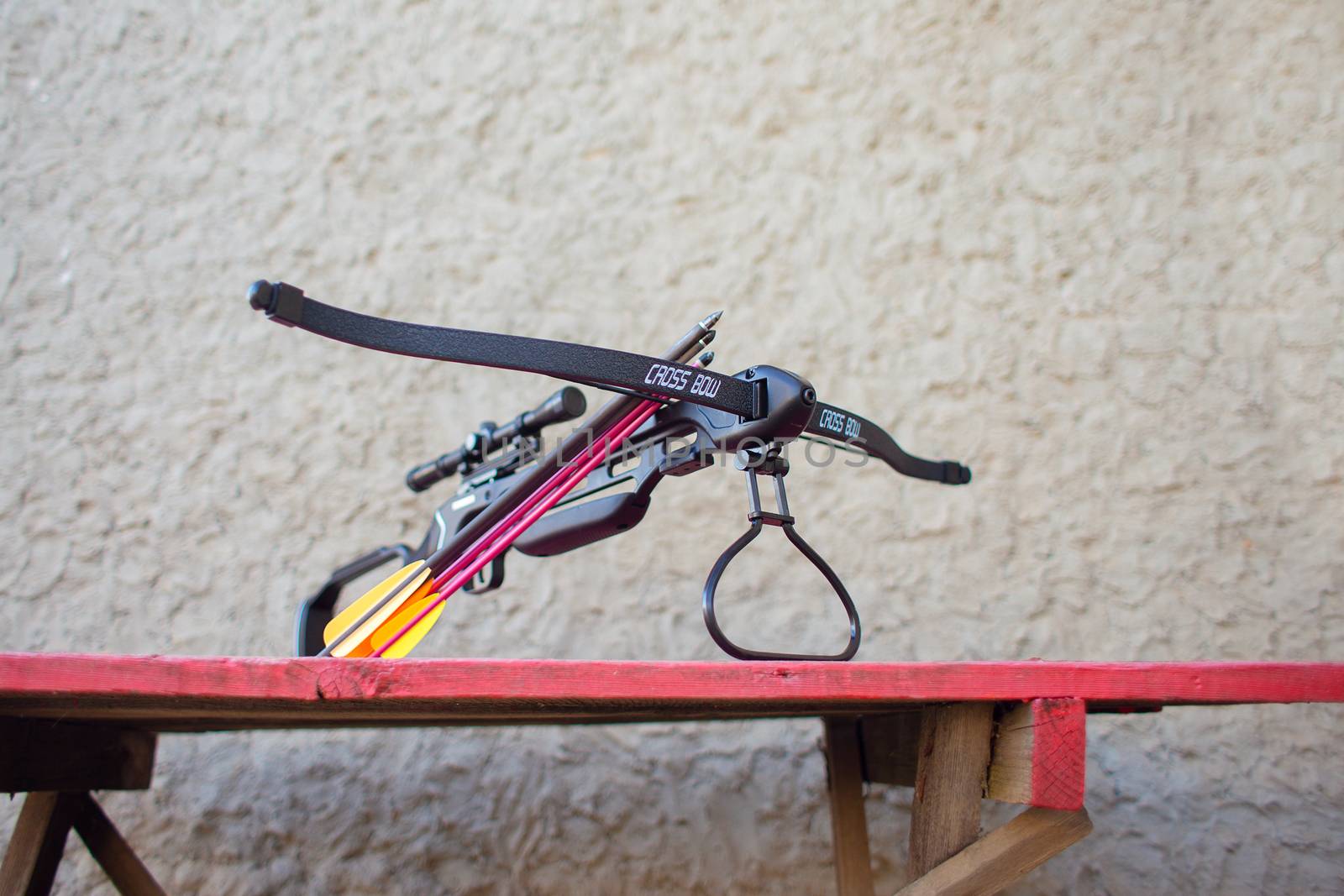 A black crossbow with a telescopic sight and arrows lies on a table in the yard. Preparing weapons for hunting