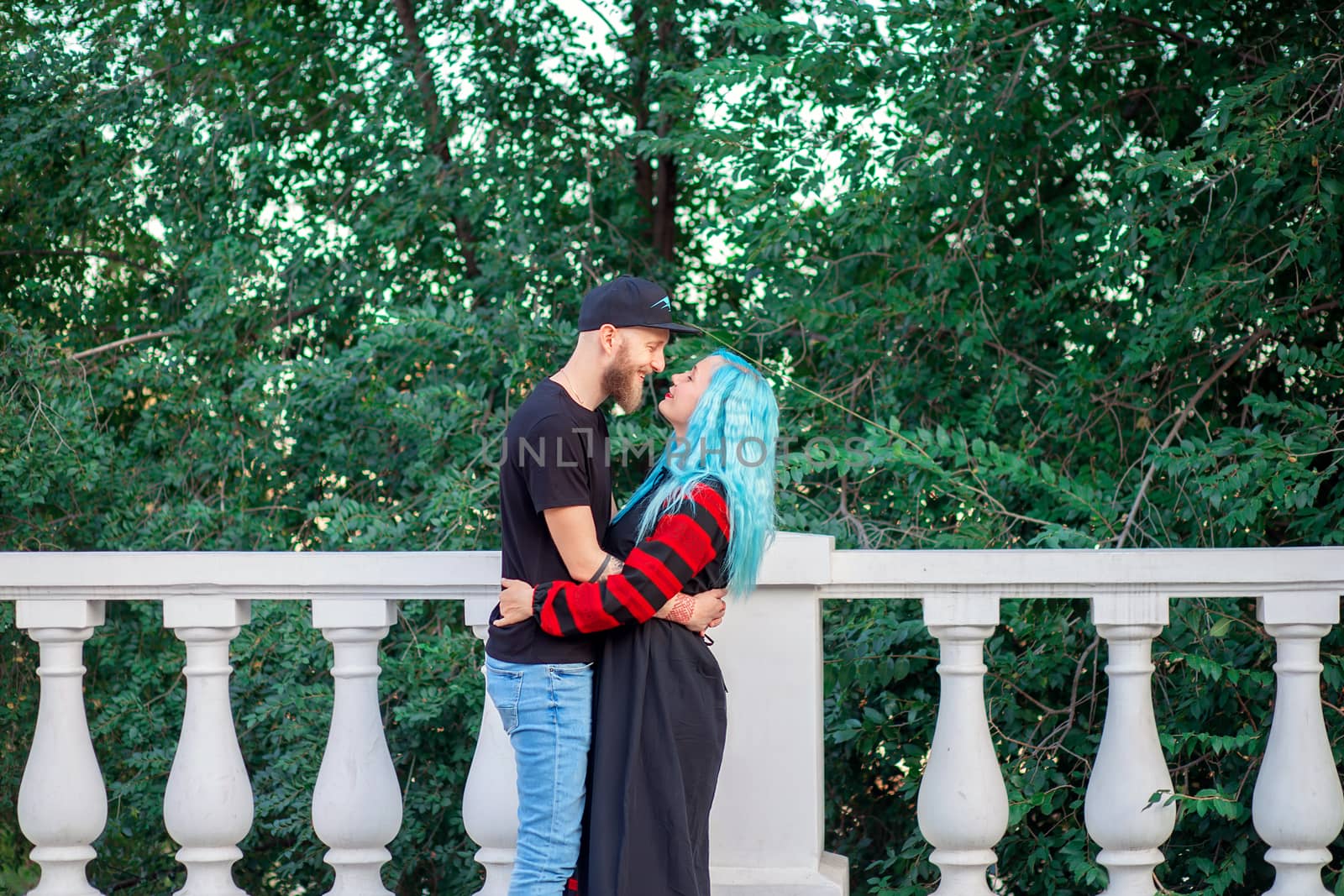 A woman with blue hair hugs her young man with a beard while walking in a city park by borisenkoket