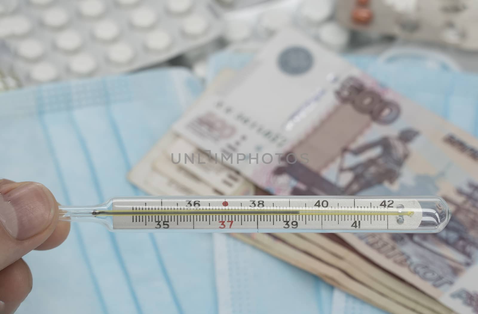 A hand holds a thermometer with a high temperature over a table with pills, a medical mask and money in rubles. Increase in prices for medicines in Russia in rubles. Russia is on quarantine