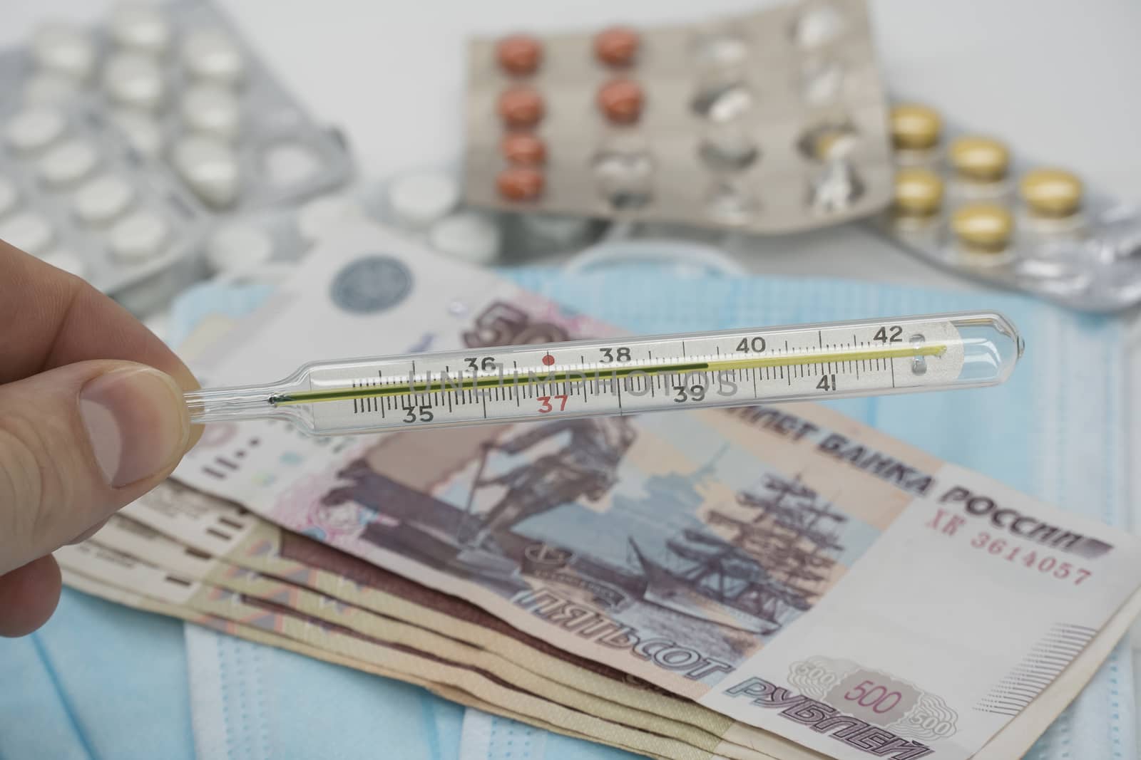 A hand holds a thermometer with a high temperature over a table with pills, a medical mask and money in rubles. Increase in prices for medicines in Russia in rubles. Russia is on quarantine