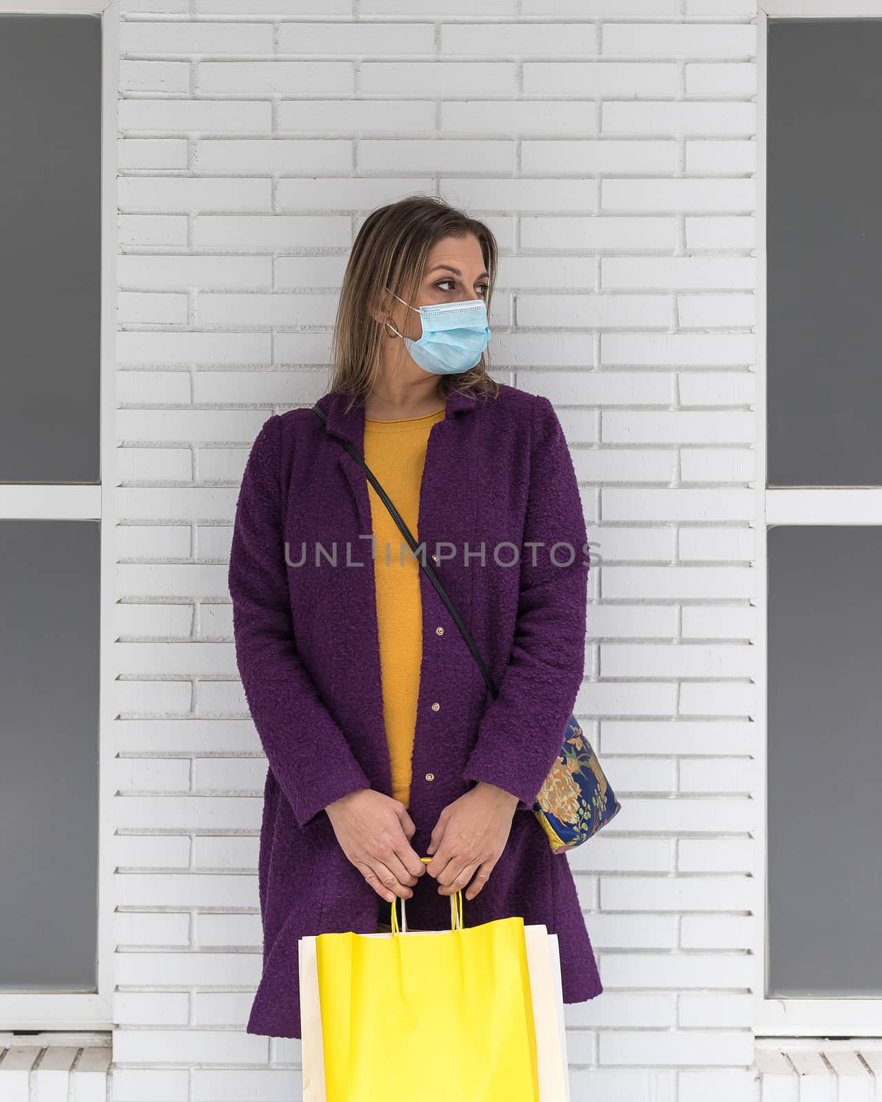 A middle-aged blonde woman wearing a surgical mask and holding shopping bags with a white brick wall in the background