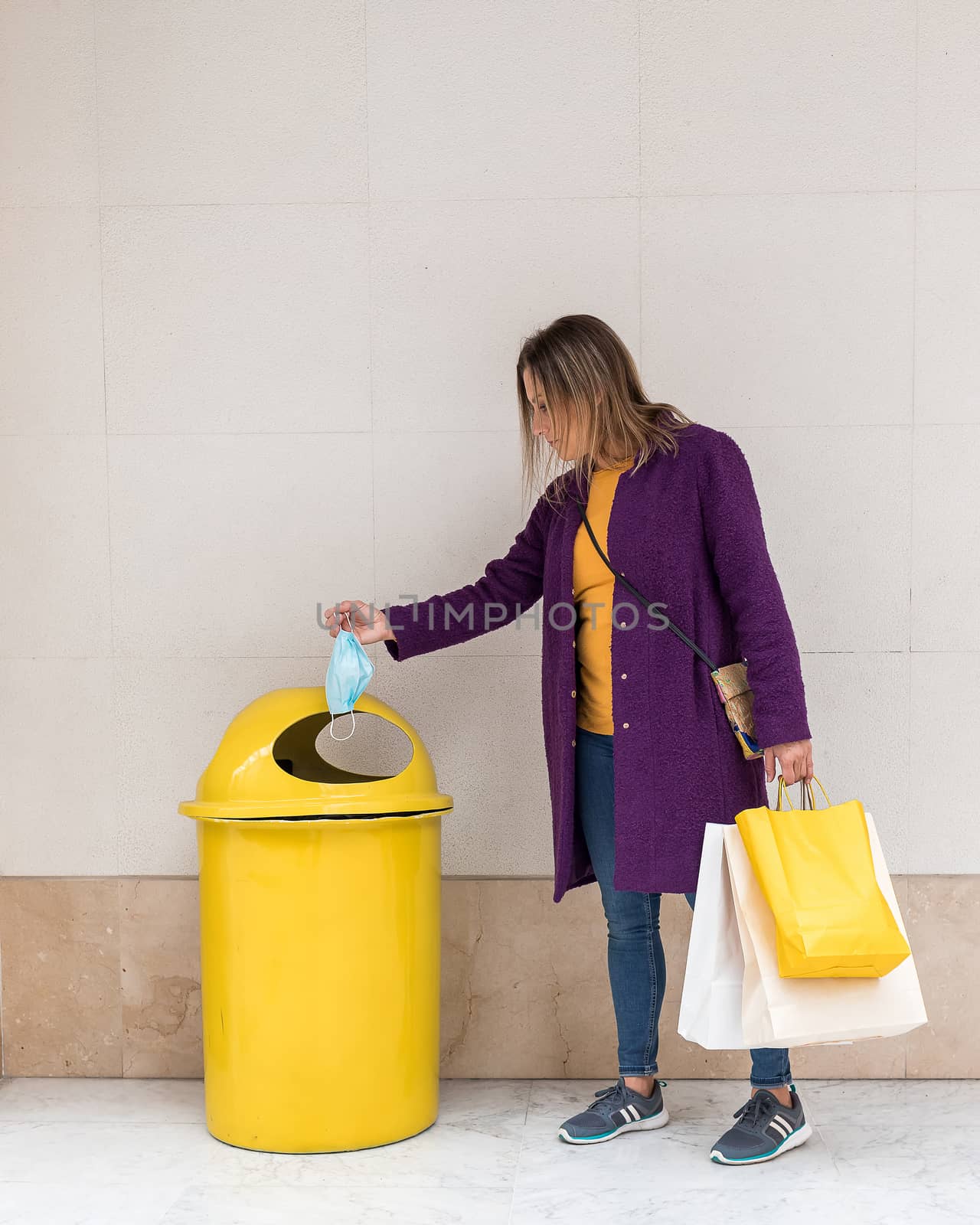 Middle-aged blonde woman throwing a face mask into a yellow garbage can while holding shopping bags