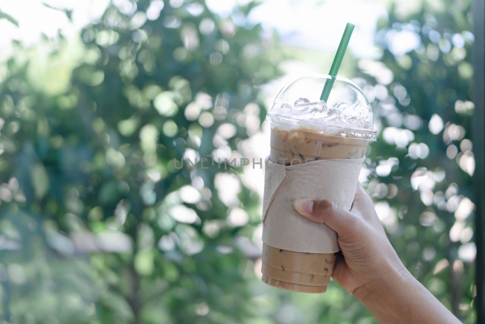 Close up hand holding glass of ice coffee with green leaves nature background, selective focus