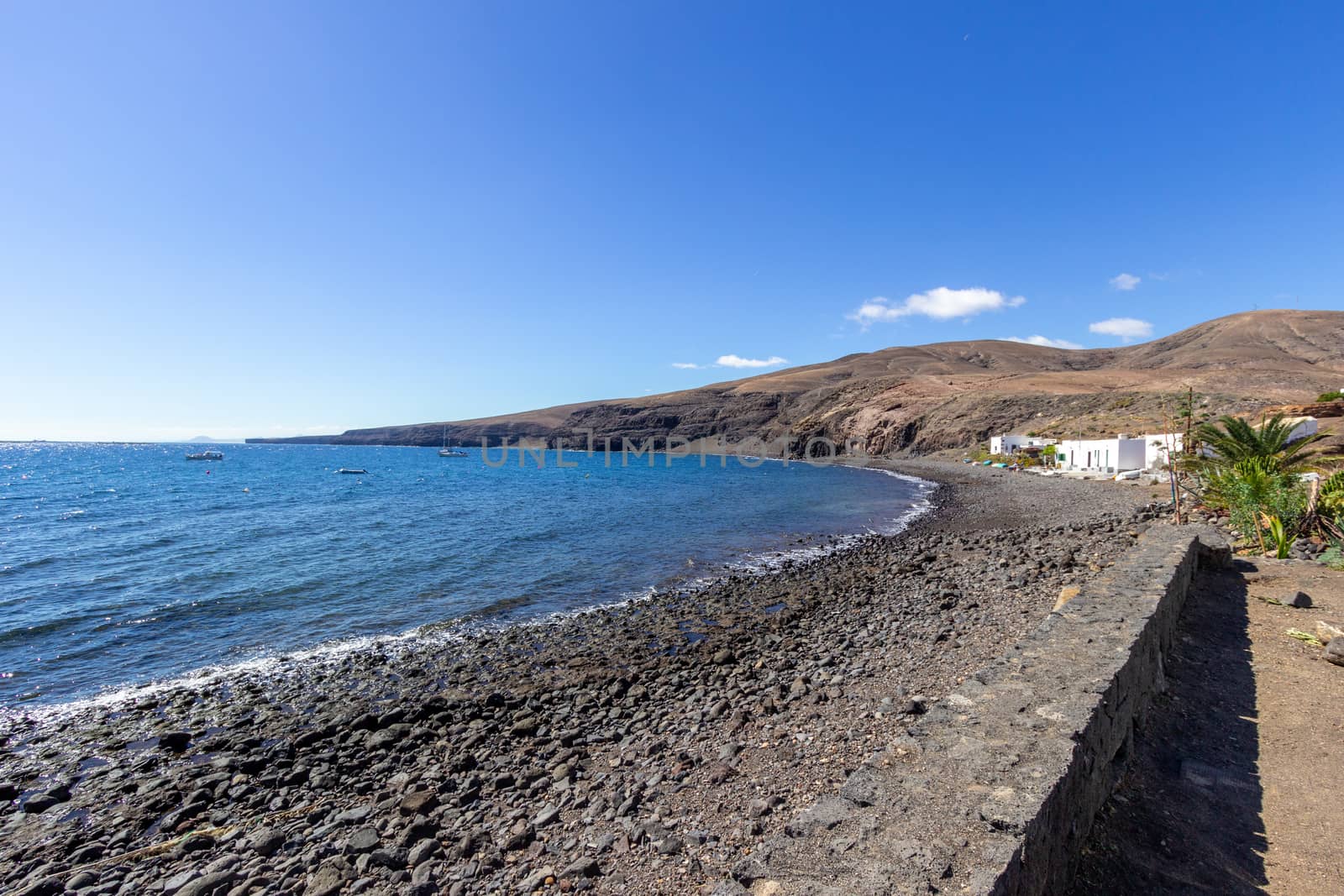 Rocky coast of Playa Quemada at Canary island Lanzarote with lava rocks, gravel beach and blue water. The sky is blue with white clouds.