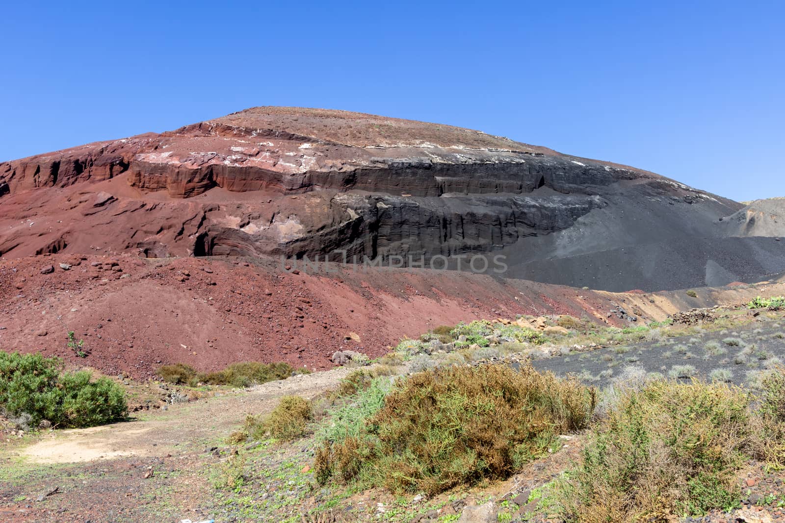 Panoramic view at red and black colored volcanic rock formation in the south of canary island Lanzarote, Spain nearby Playa Blanca with sparse vegetation in the foreground and blue sky