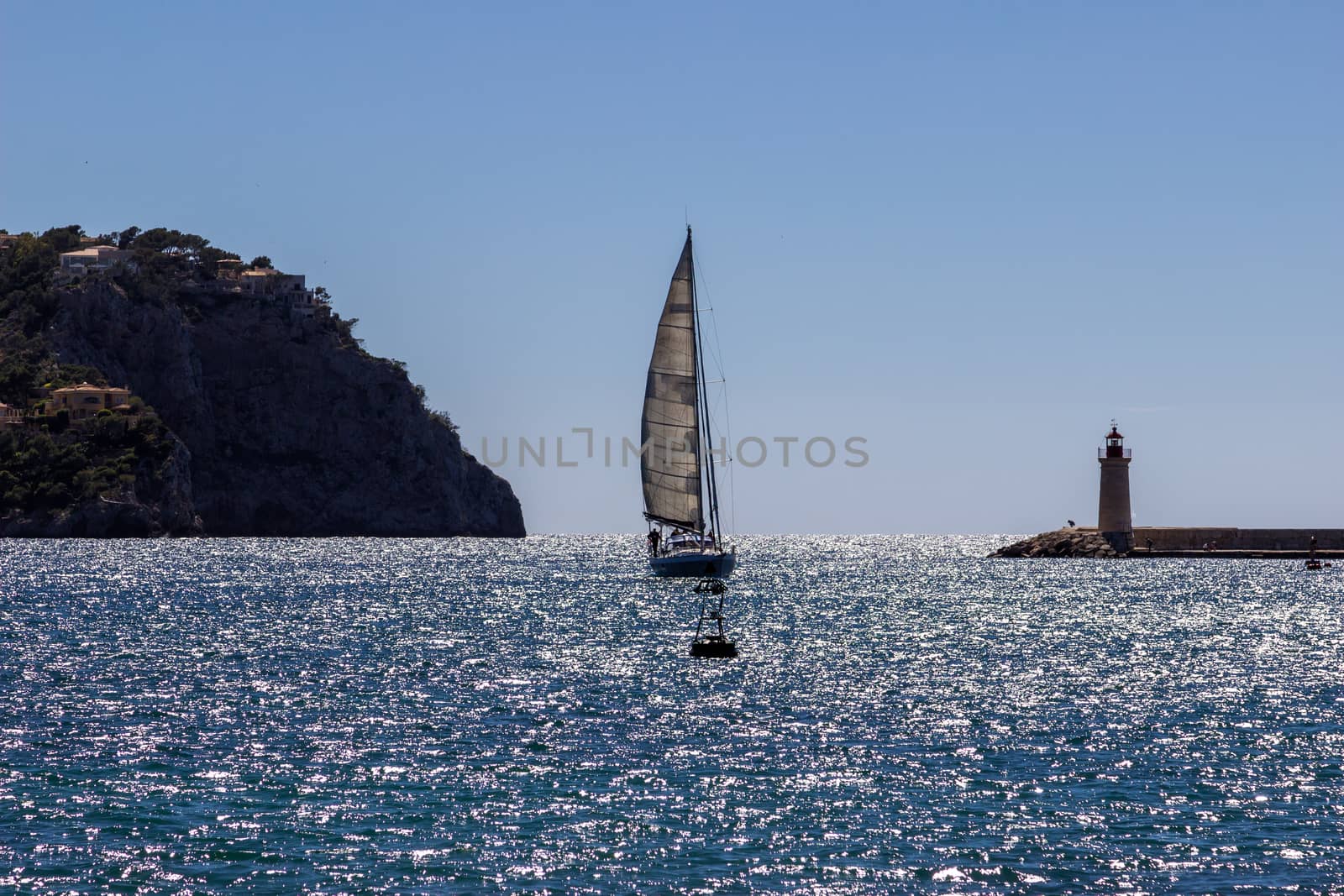  Sailboat in the harbor entrance of Port d' Andratx between a Lighthouse and a mountain with houses