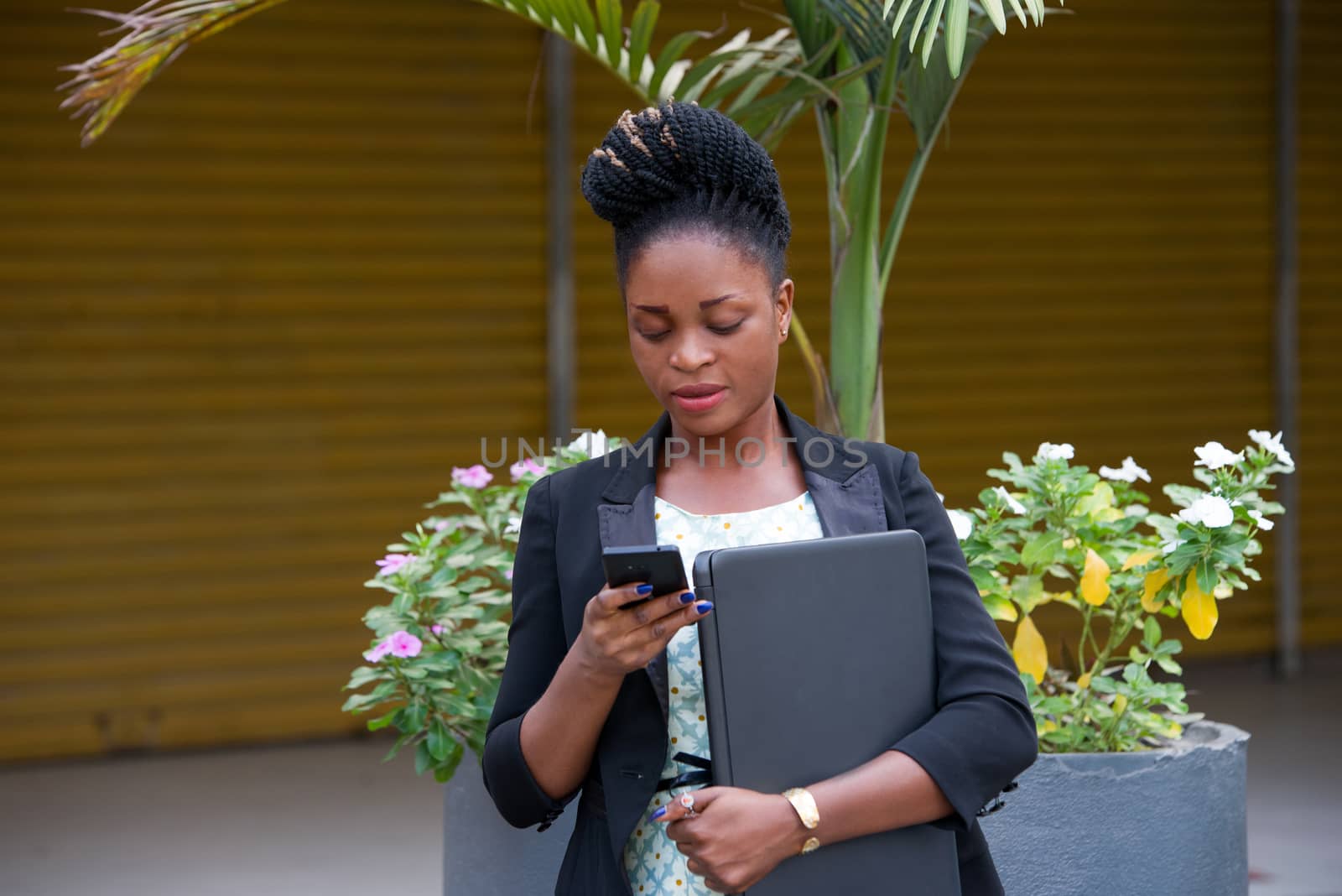 young businesswoman standing with a laptop, reading messages in her mobile phone.