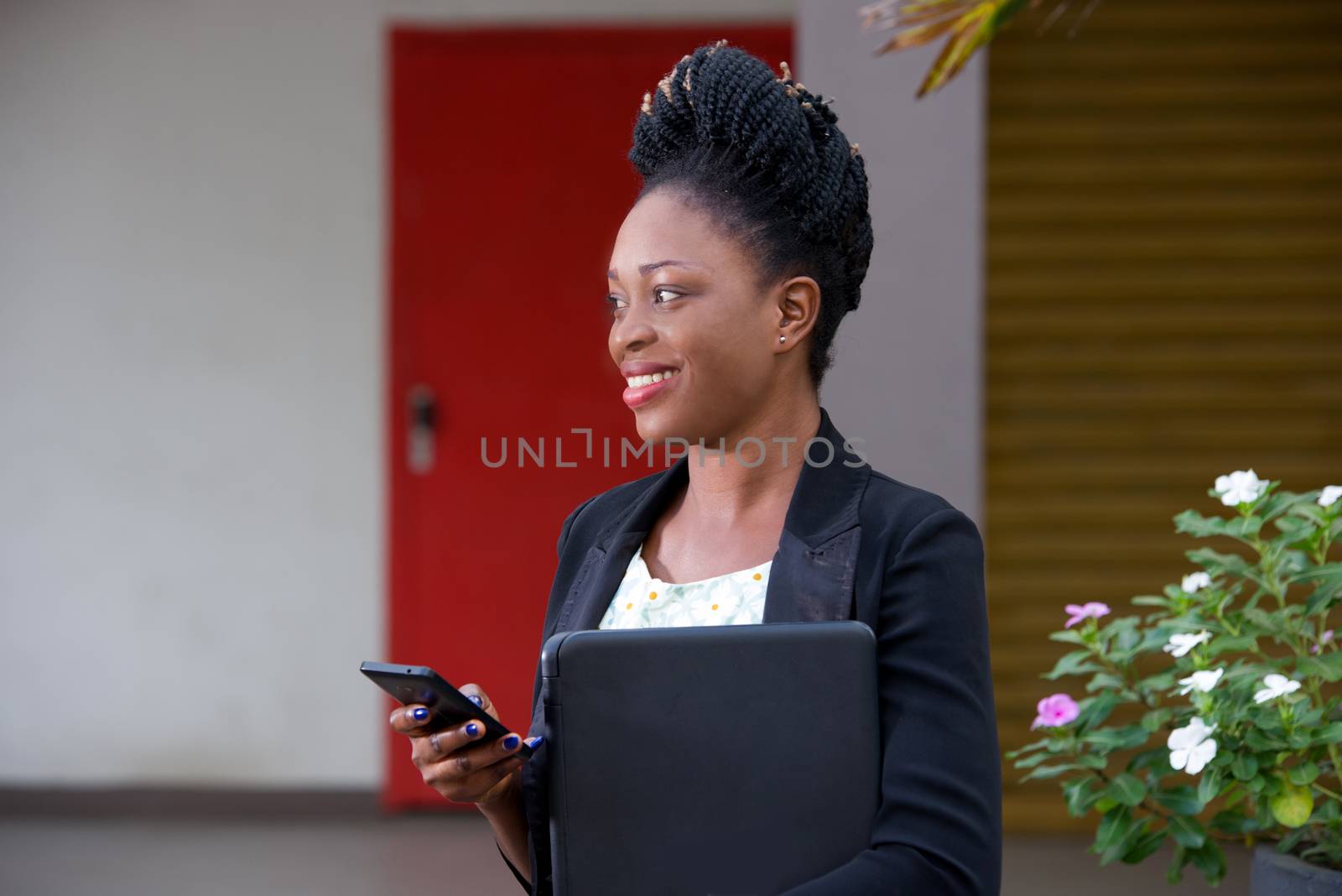 Smiling businesswoman using smart phone while standing outside holding a laptop.