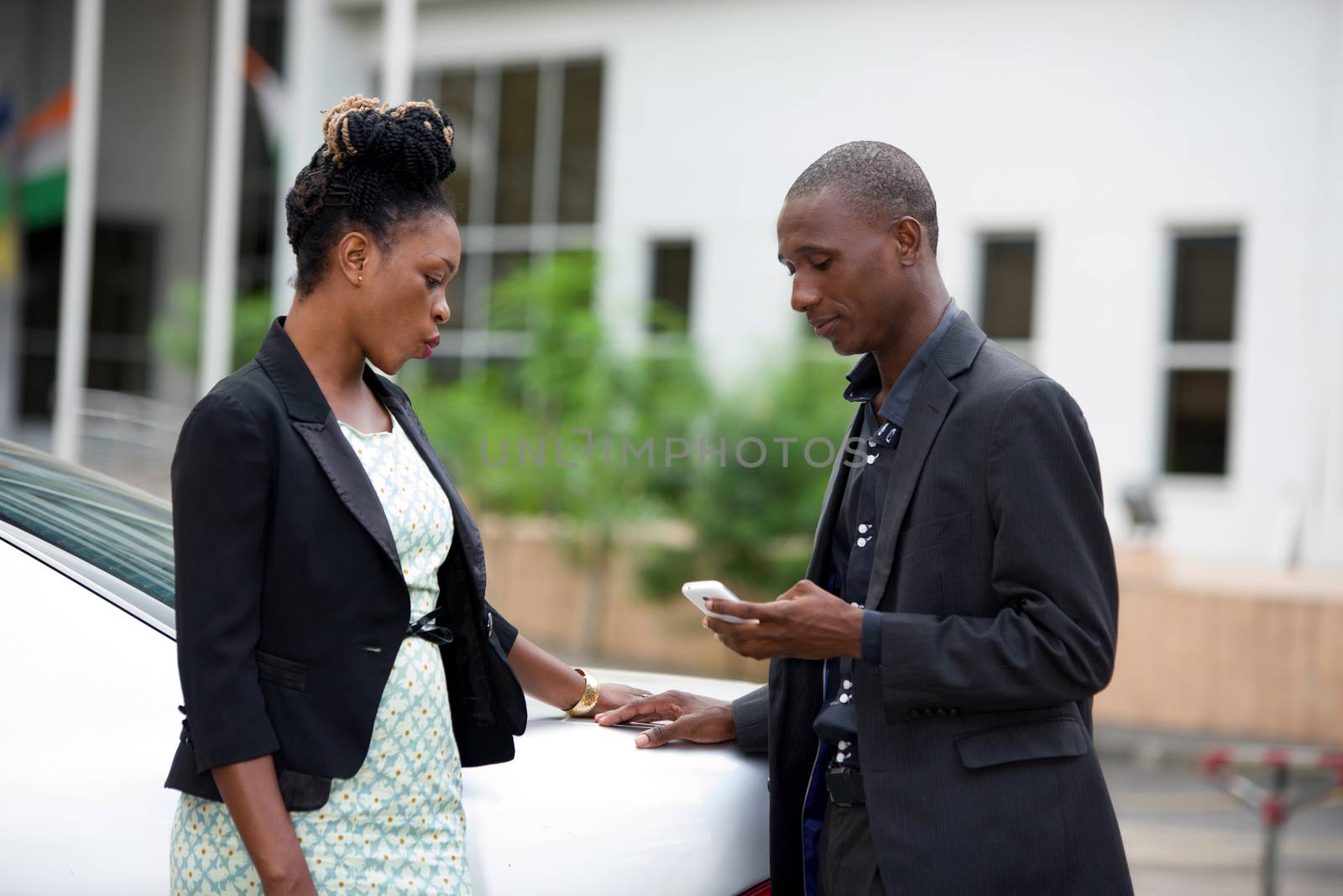 Portrait of middle-aged business man and woman relaxing and chatting outside beside a car with office building in the background. The man is using a smart phone.