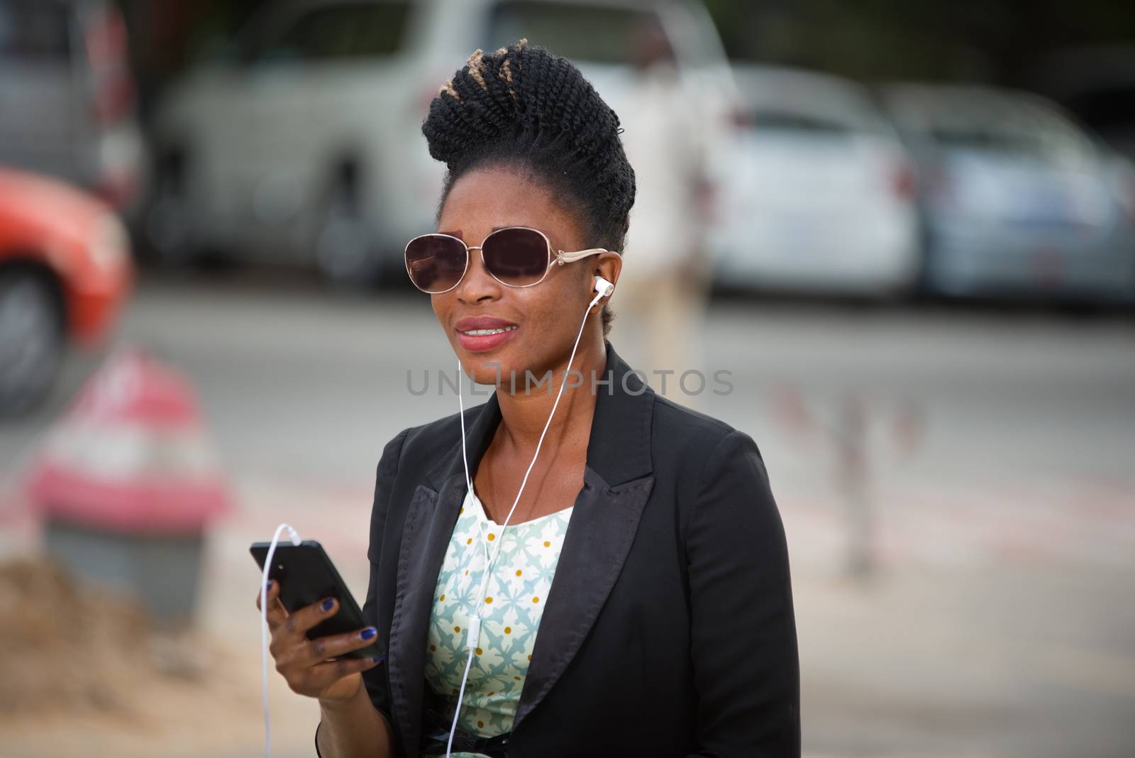 young woman wearing a black jacket and sunglasses sits outside and listens to music with her mobile phone