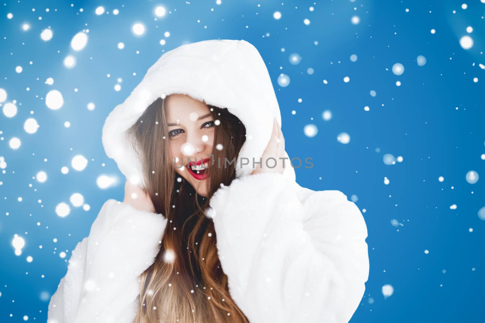 Happy Christmas and winter holiday portrait of young woman in white hooded fur coat, snow on blue background, fashion and lifestyle campaign