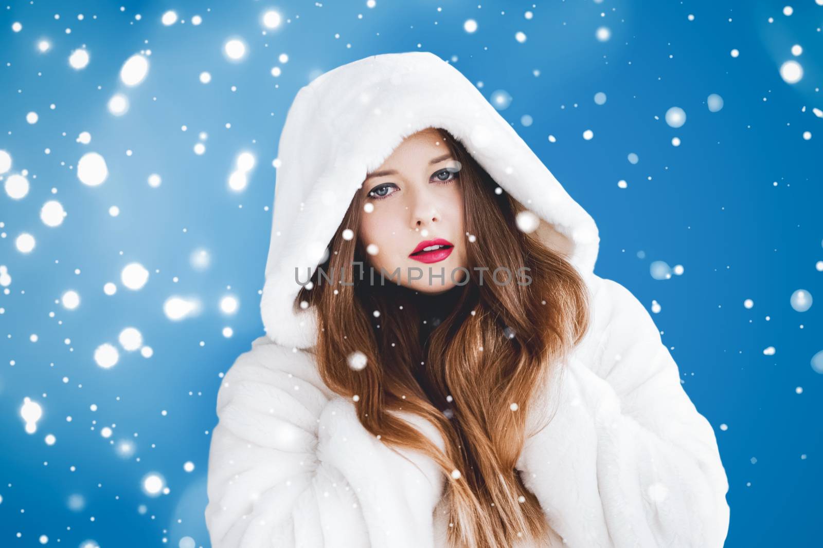 Happy Christmas and winter holiday portrait of young woman in white hooded fur coat, snow on blue background, fashion and lifestyle campaign
