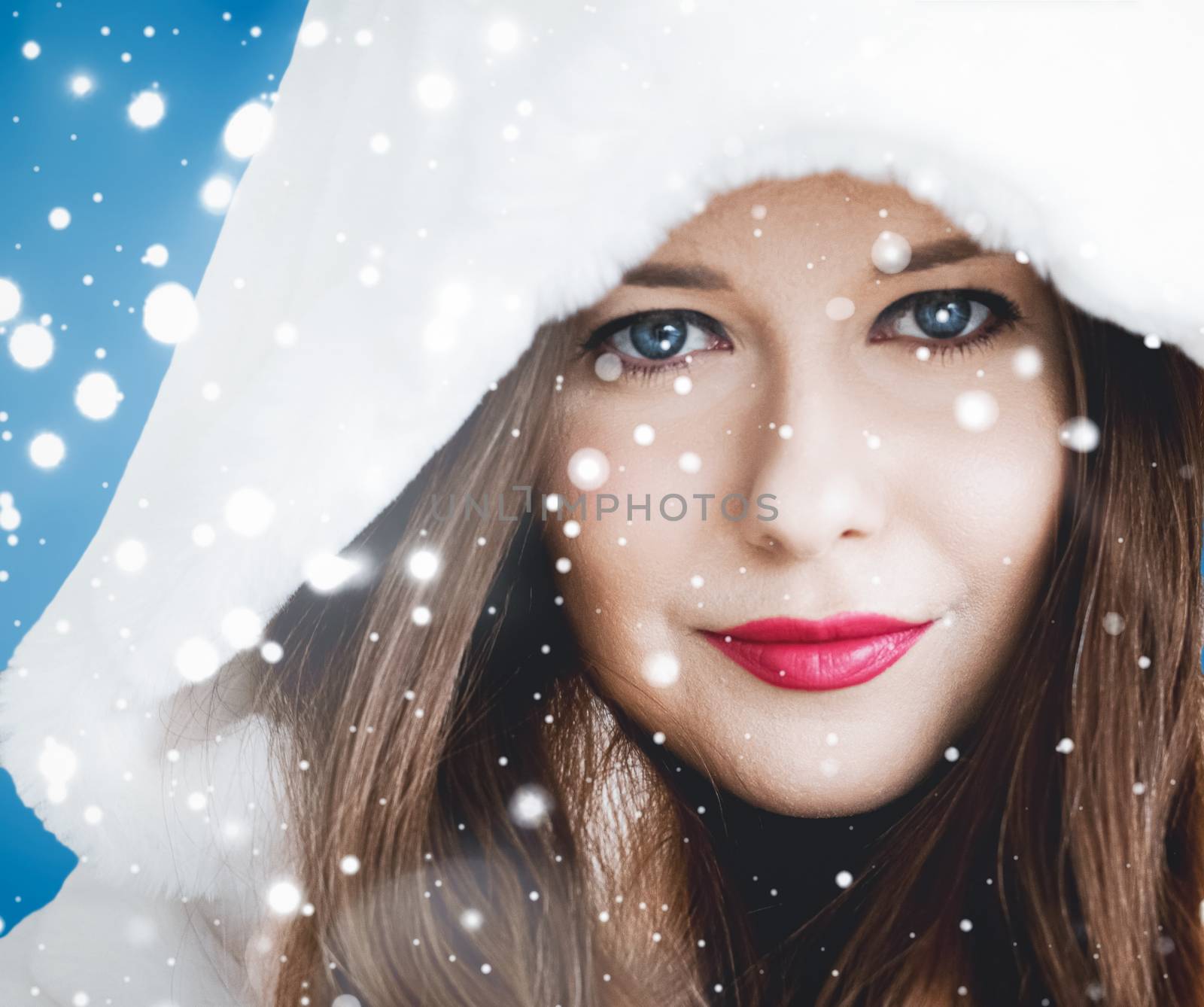 Happy Christmas and winter holiday portrait of young woman in white hooded fur coat, snow on blue background, fashion and lifestyle campaign