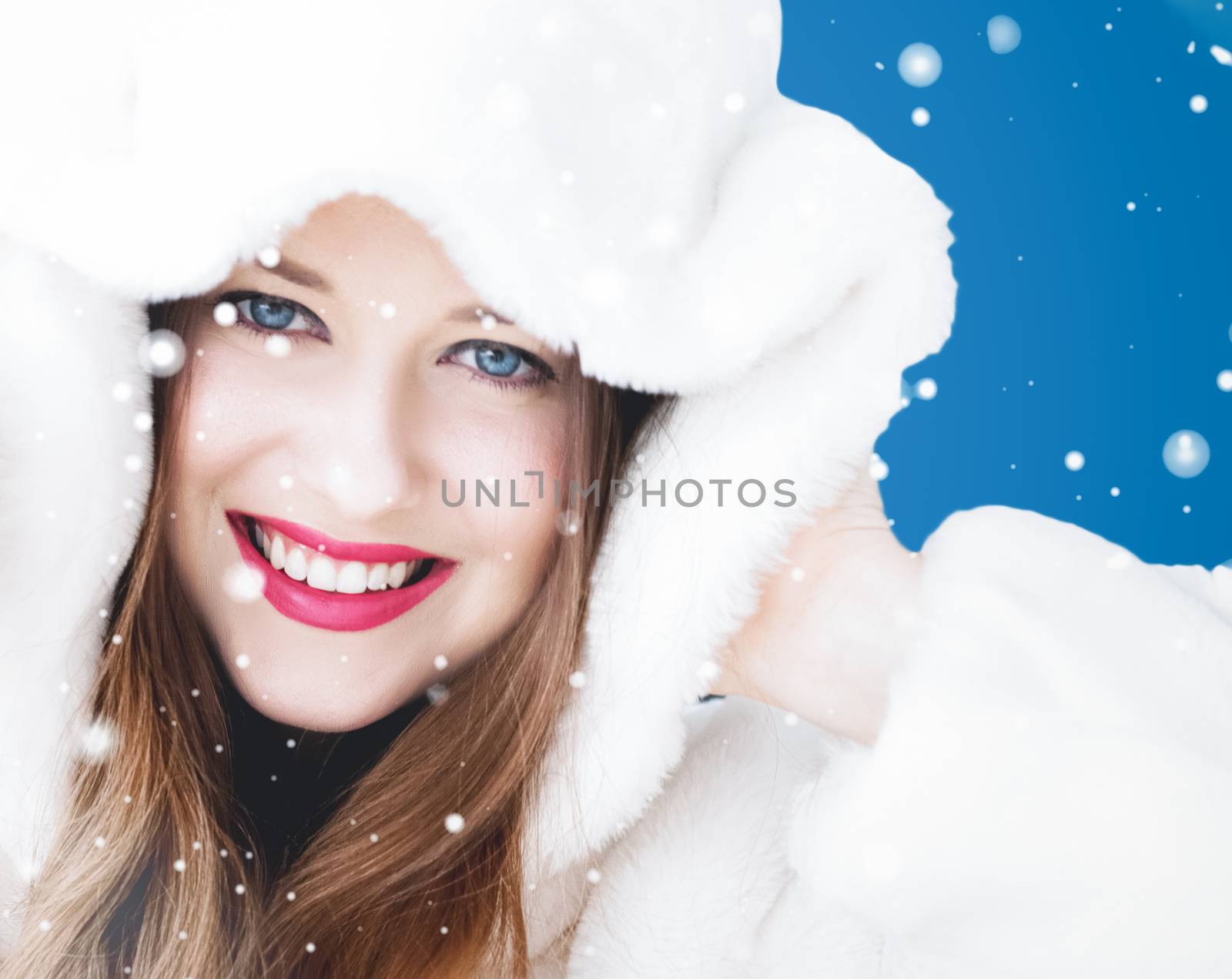 Happy Christmas and winter holiday portrait of young woman in white hooded fur coat, snow on blue background, fashion and lifestyle campaign