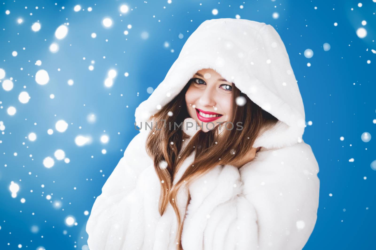 Happy Christmas and winter holiday portrait of young woman in white hooded fur coat, snow on blue background, fashion and lifestyle campaign