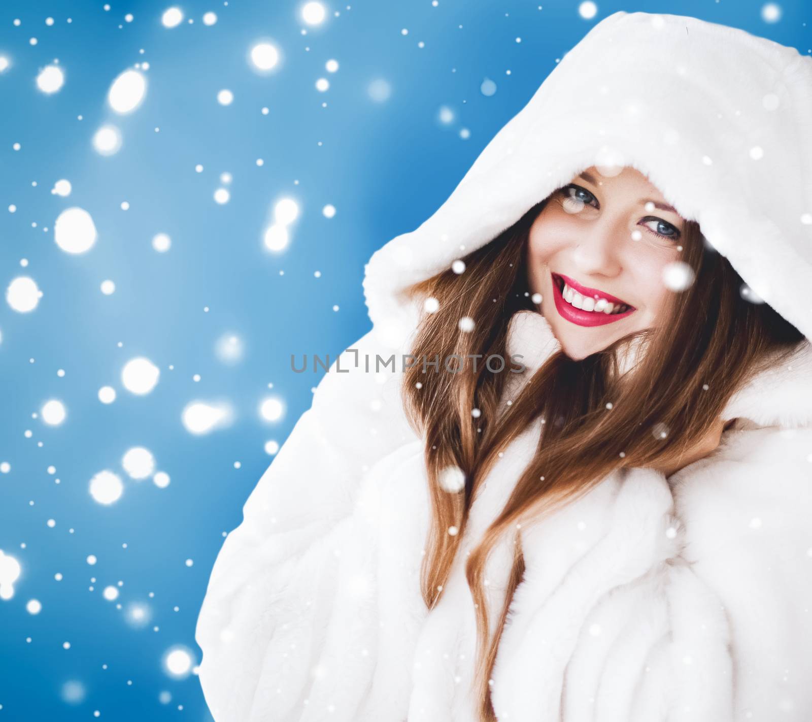 Happy Christmas and winter holiday portrait of young woman in white hooded fur coat, snow on blue background, fashion and lifestyle campaign