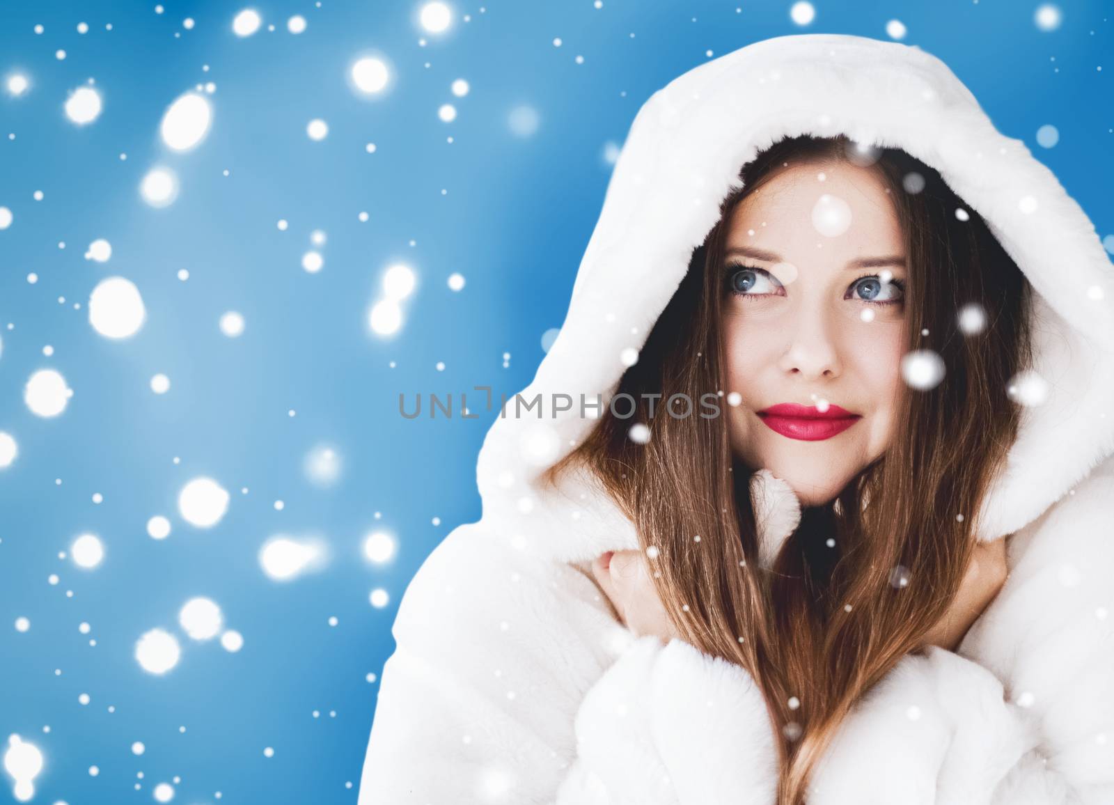 Happy Christmas and winter holiday portrait of young woman in white hooded fur coat, snow on blue background, fashion and lifestyle campaign