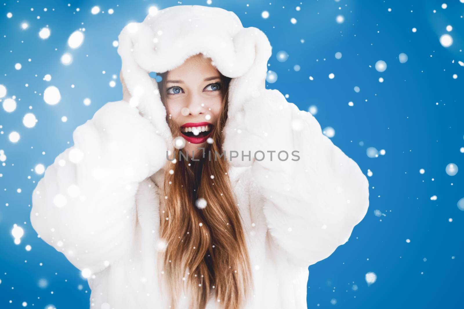 Happy Christmas and winter holiday portrait of young woman in white hooded fur coat, snow on blue background, fashion and lifestyle campaign