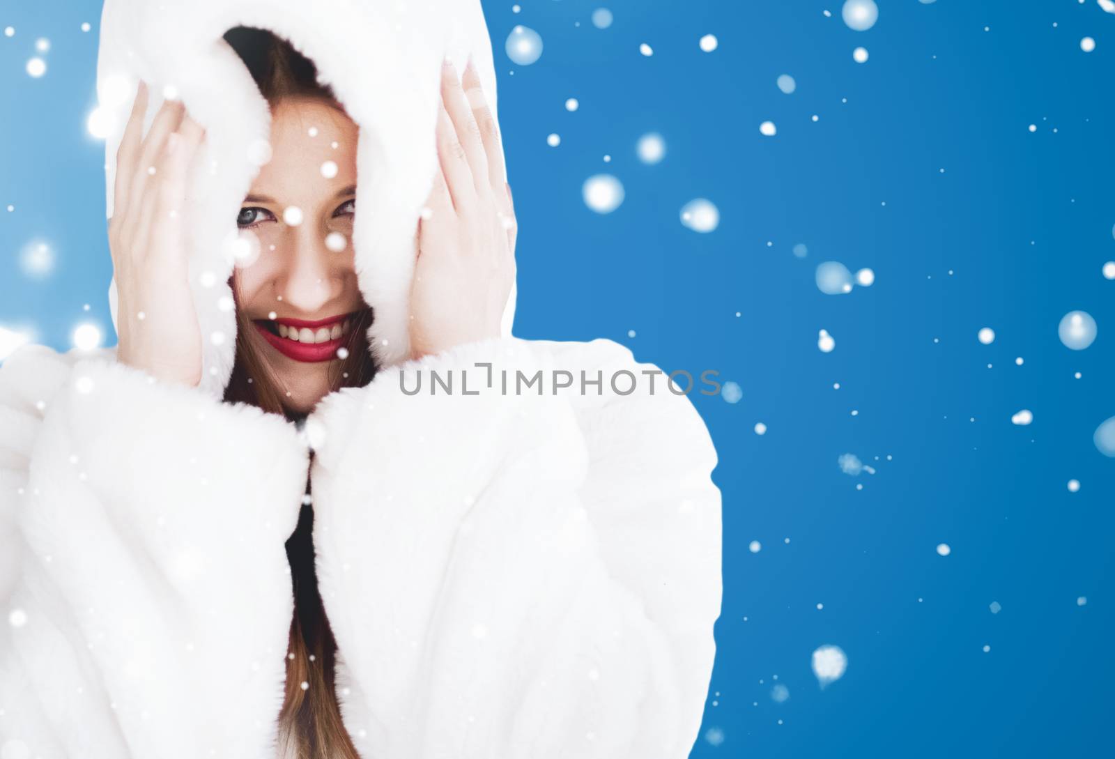 Happy Christmas and winter holiday portrait of young woman in white hooded fur coat, snow on blue background, fashion and lifestyle campaign