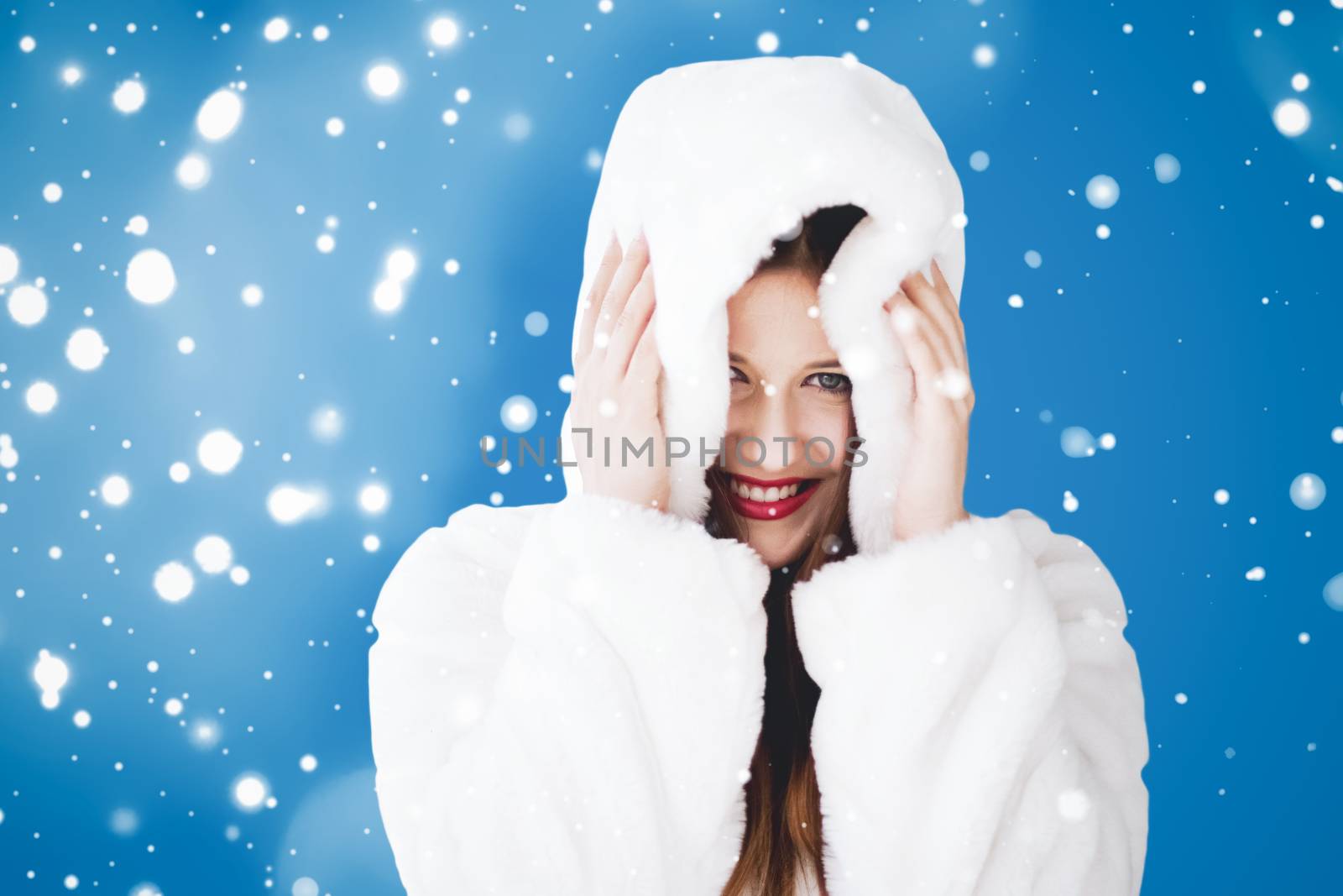 Happy Christmas and winter holiday portrait of young woman in white hooded fur coat, snow on blue background, fashion and lifestyle campaign