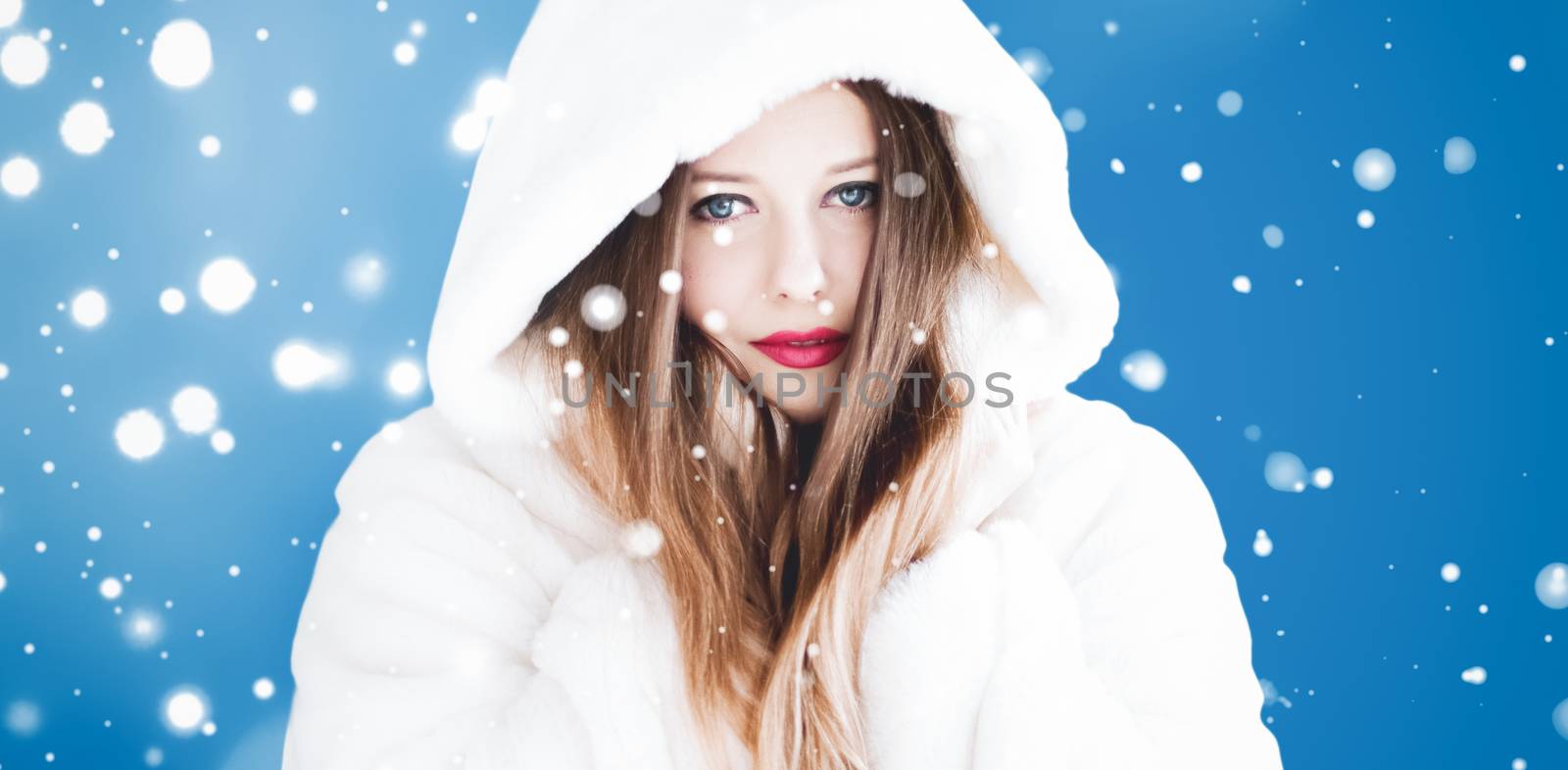 Happy Christmas and winter holiday portrait of young woman in white hooded fur coat, snow on blue background, fashion and lifestyle campaign