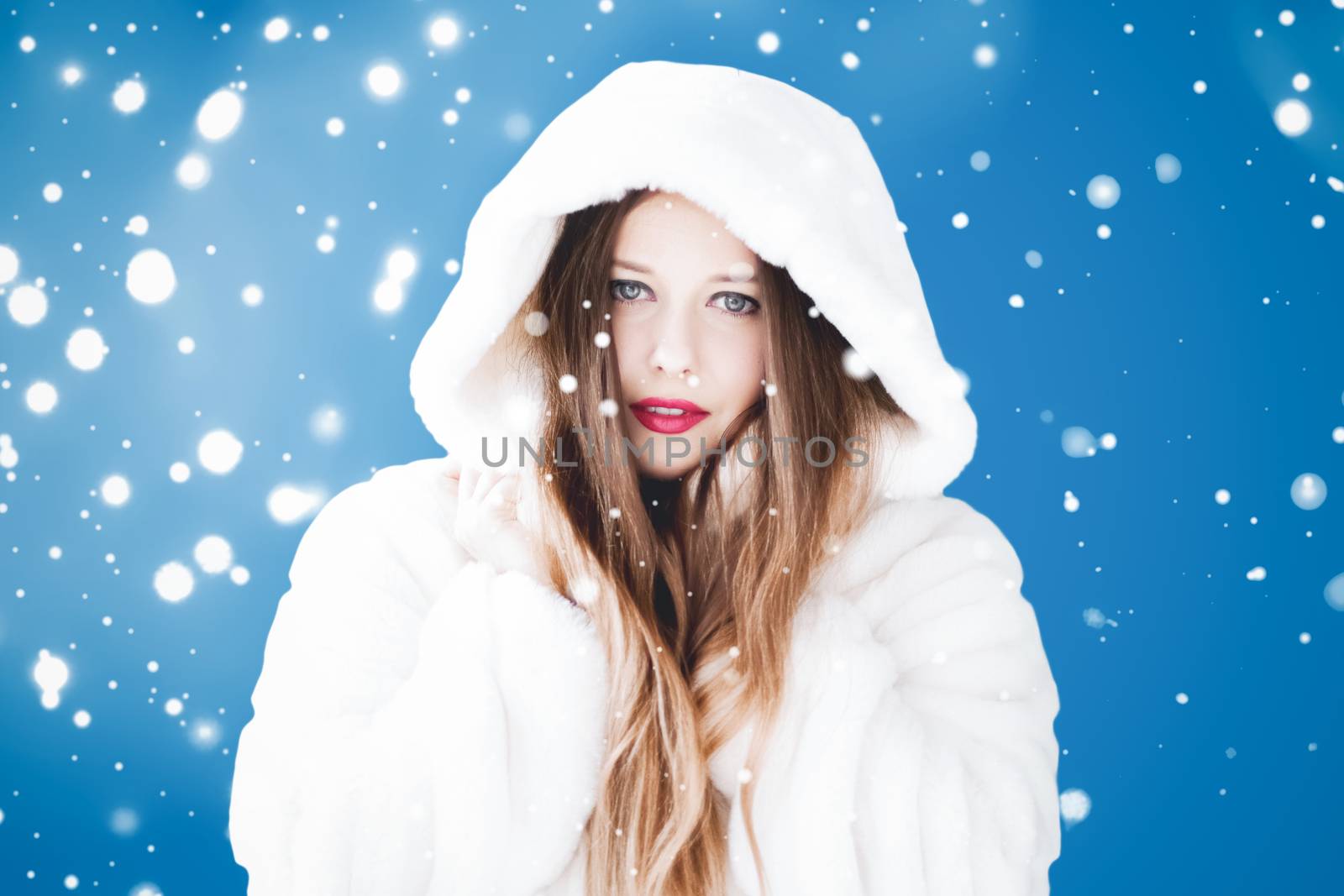 Happy Christmas and winter holiday portrait of young woman in white hooded fur coat, snow on blue background, fashion and lifestyle campaign