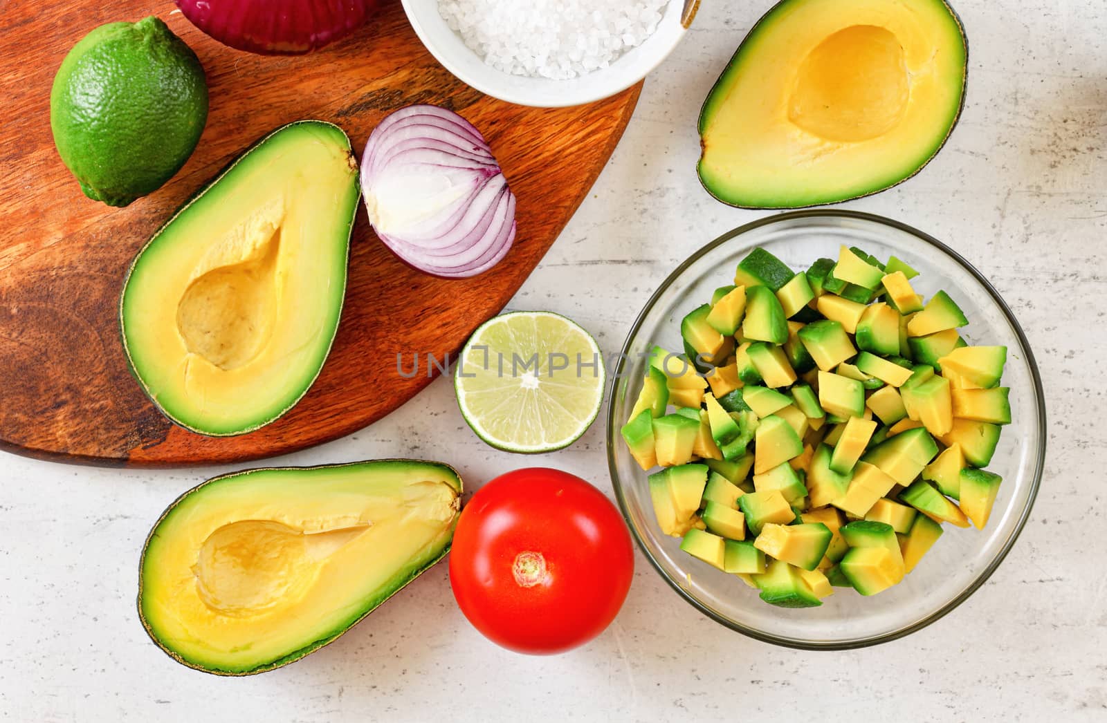 Avocado halves, some cut to small pieces, limes, tomato and onion - basic guacamole ingredients on white working board, view from above by Ivanko