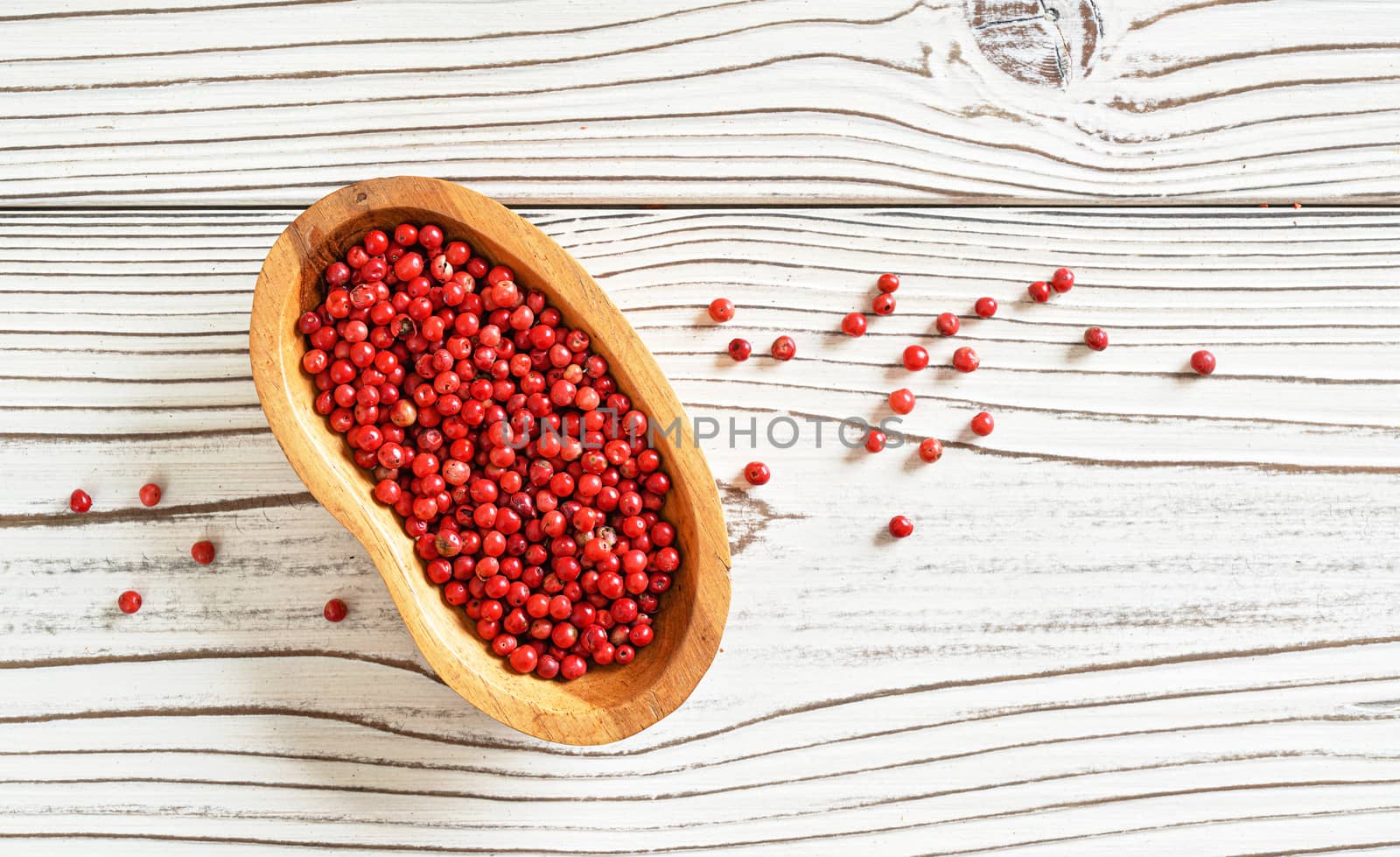 Red or Pink peppercorn in small wooden bowl, some scattered on white boards desk, closeup photo from above by Ivanko