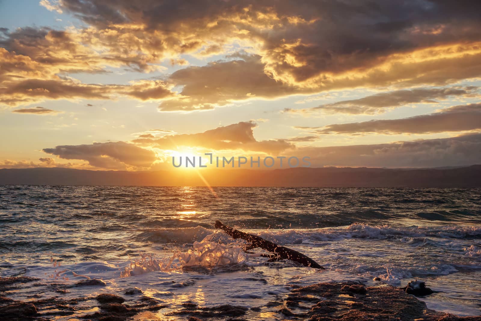 Sun shines through dramatic sunset clouds to waves at Jordanian side of Dead Sea.