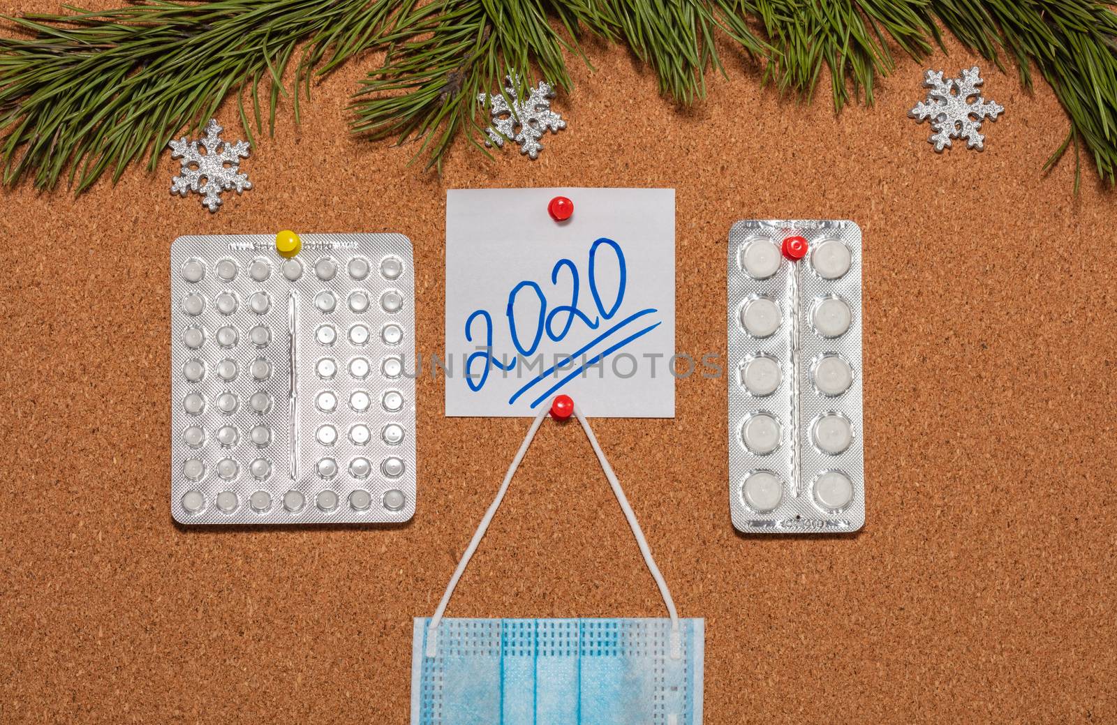 Medical mask, white pills, white sticky note with 2020 pinned on notice board which is decorated with pine twigs and snowflakes. Healthcare, christmas, new years celebration, new normal concepts.