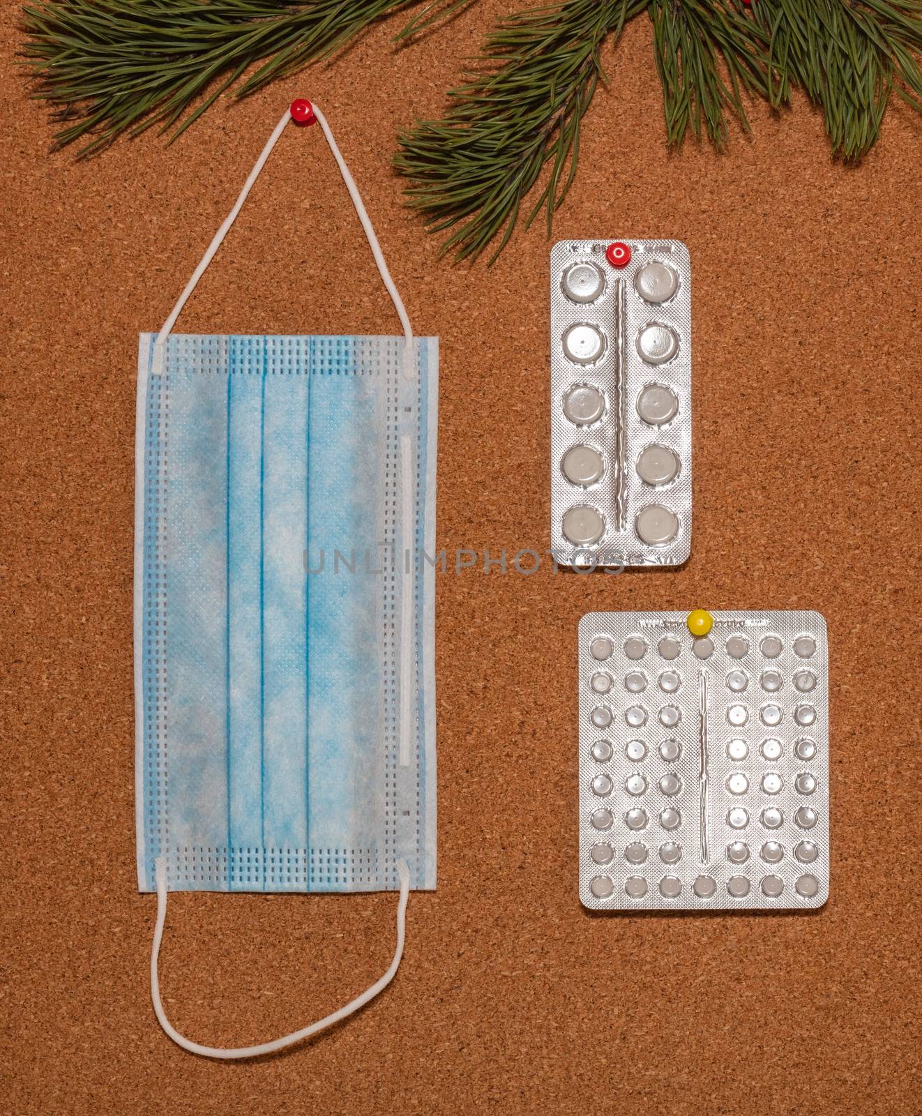 Medical mask and white pills pinned on a corkwood notice board which is decorated with pine twigs at the top. Healthcare, christmas, new years celebration, new normal concepts. by DamantisZ