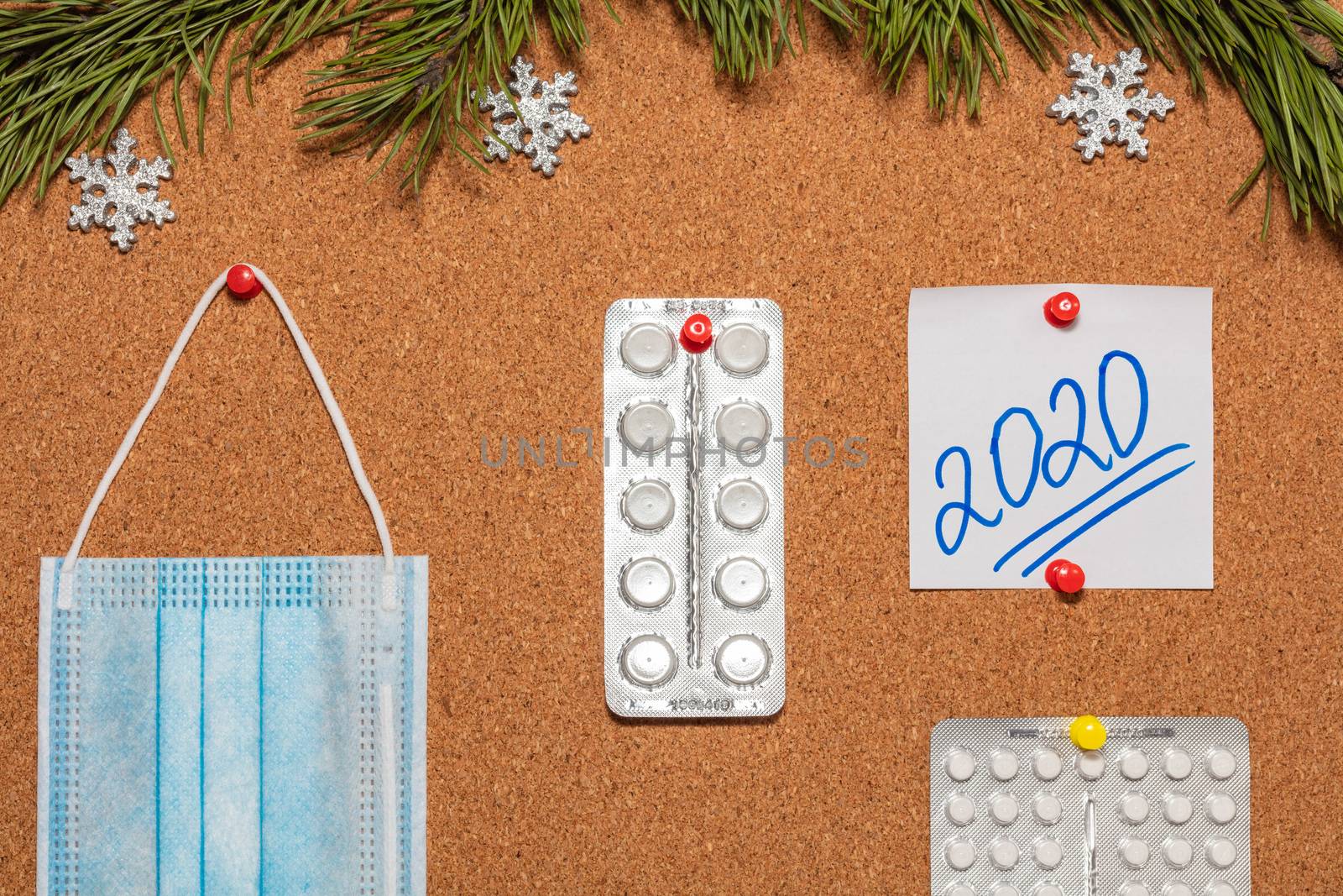 Medical mask, white pills, white sticky note with 2020 pinned on notice board which is decorated with pine twigs and snowflakes. Healthcare, christmas, new years celebration, new normal concepts. by DamantisZ